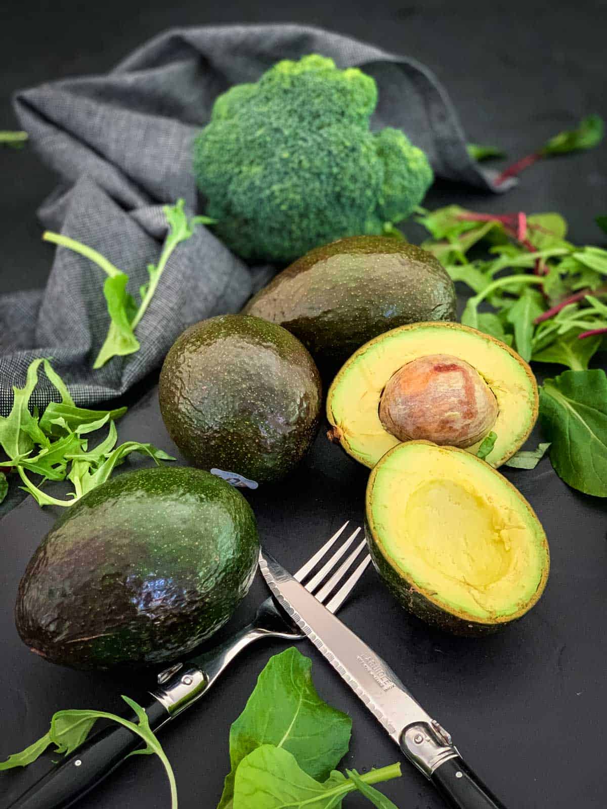 Avocados whole and half against a backdrop of a blue napkin, broccoli and mixed leaves