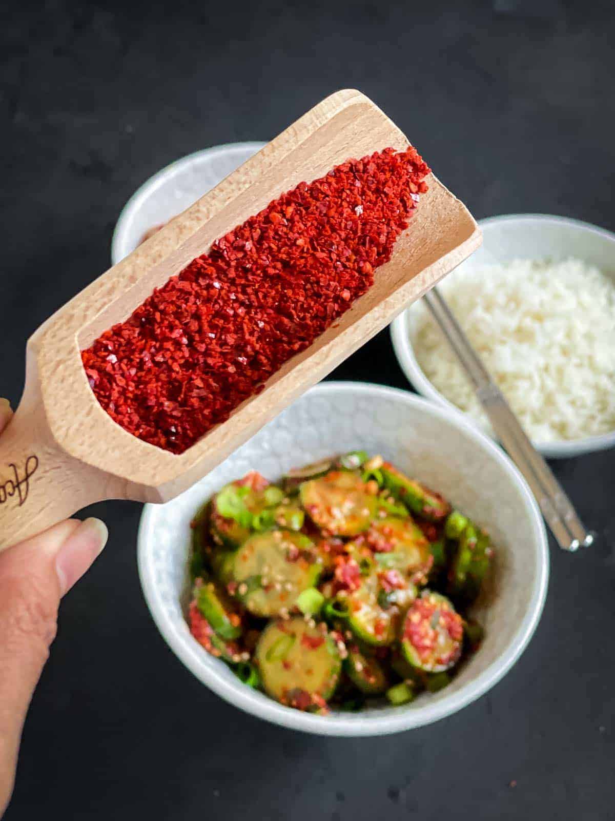 Gochugaru - Korean red chilli flakes, in a wooden spoon over cucumber salad and rice