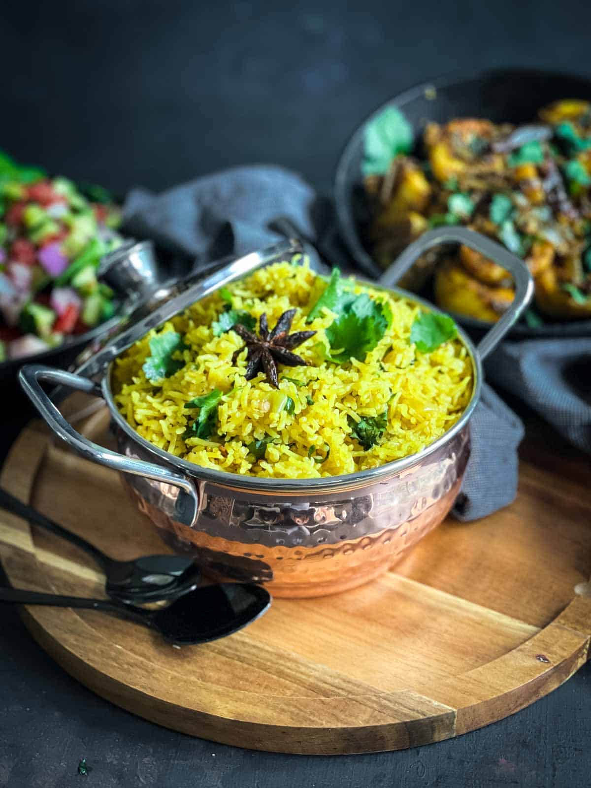 Turmeric Rice in a copper kadai on a wooden board served with kachumber salad and Bombay aloo