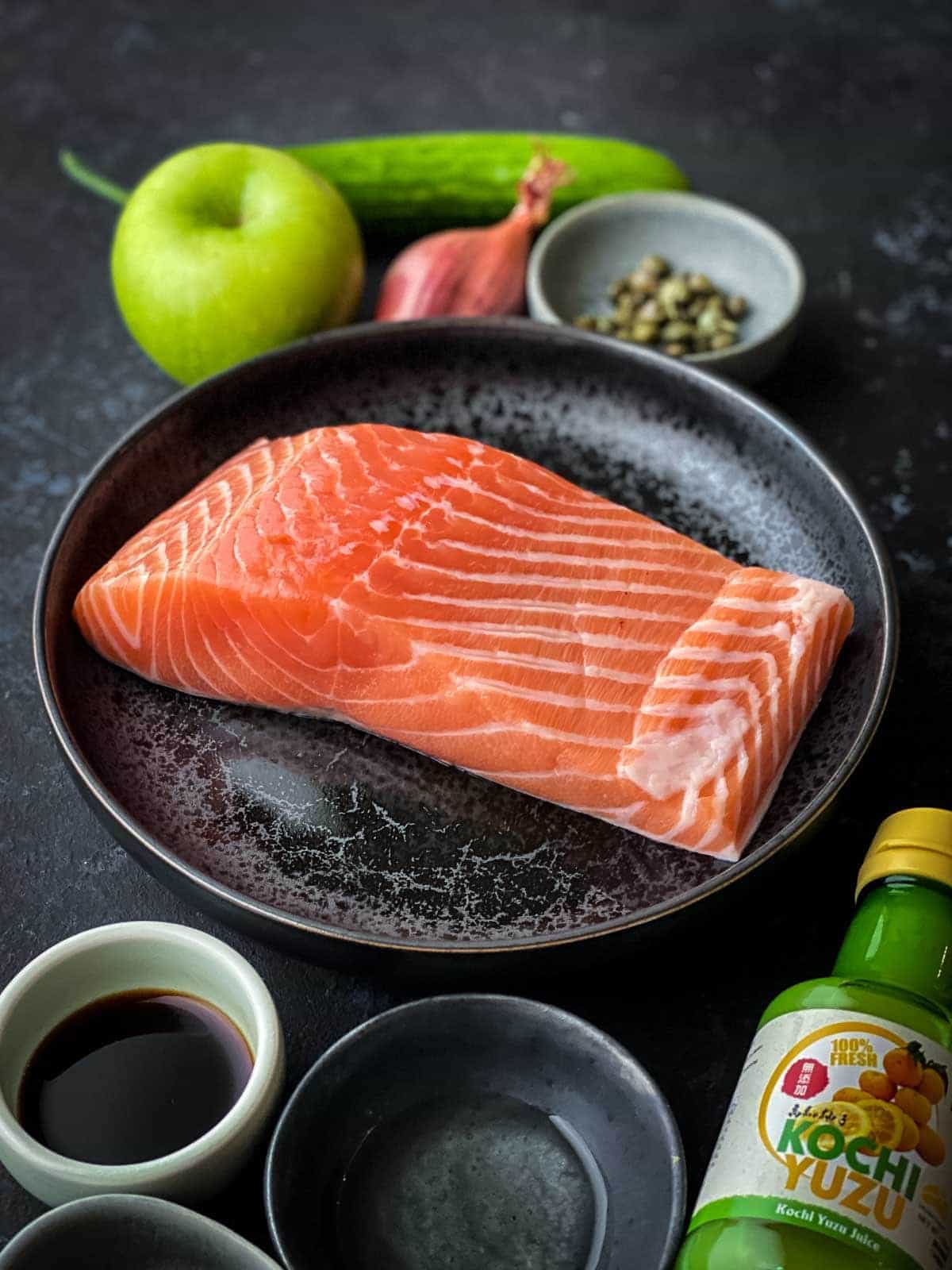 Raw sashimi grade salmon on a black speckled plate surrounded by the rest of the ingredients of the recipe