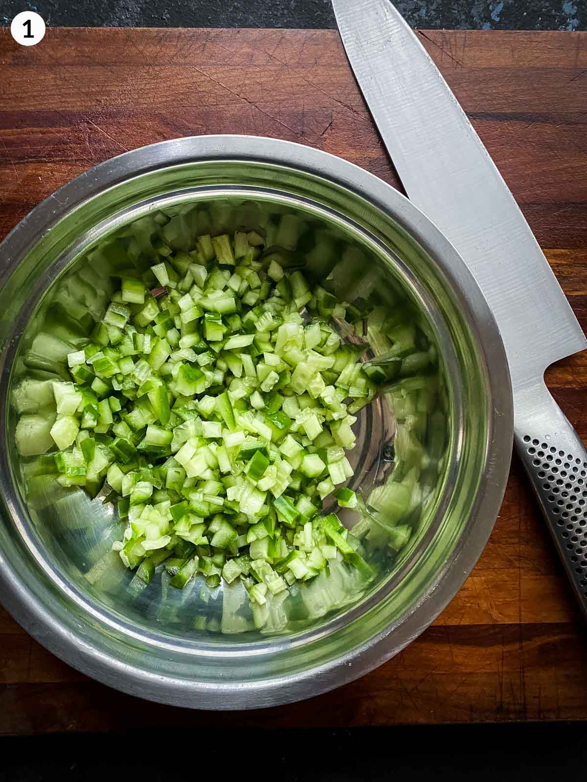 Diced cucumber in a prep bowl on a wooden board with a sharp knife