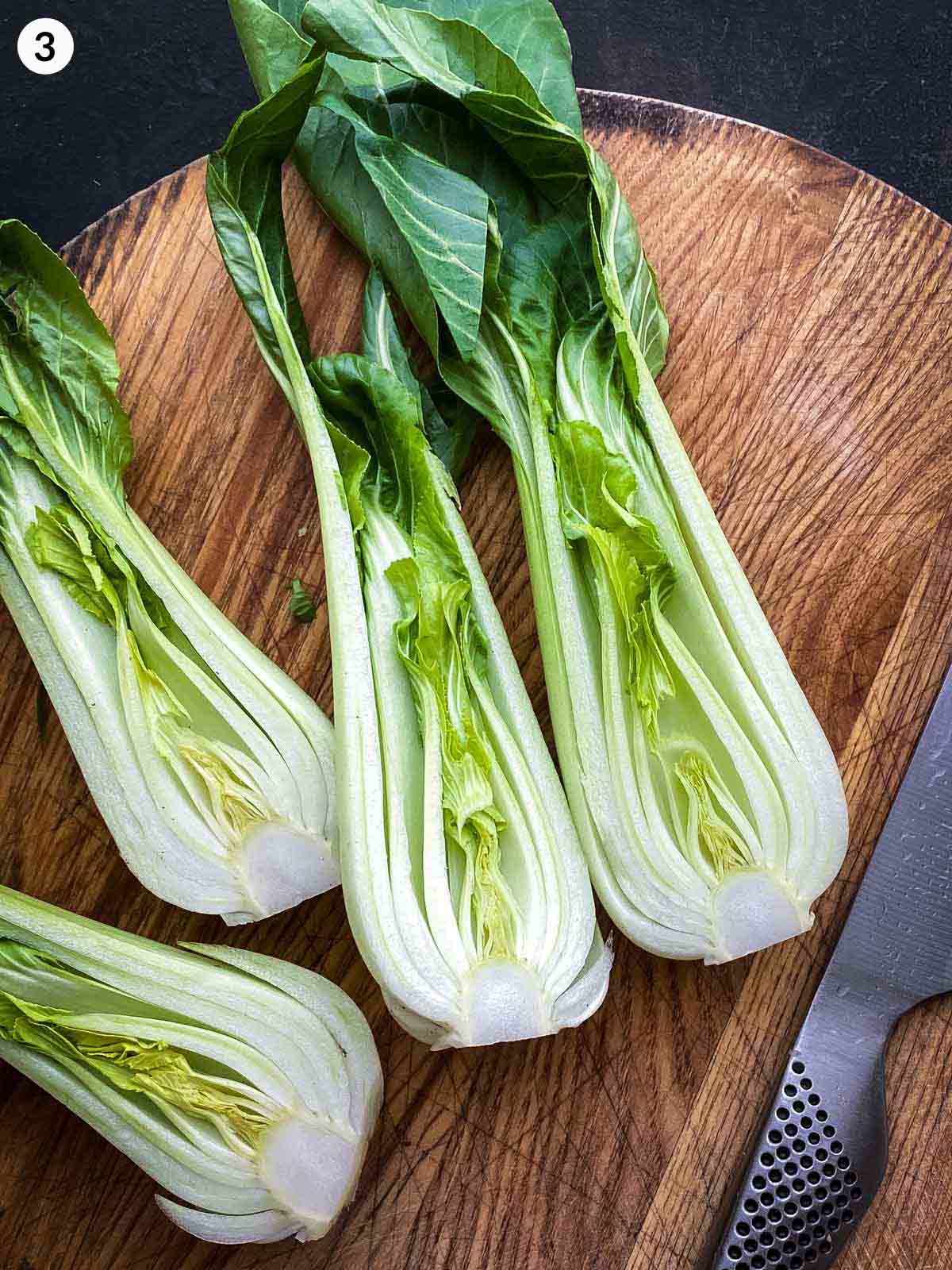 Cutting bok choy in half with a knife resting a on a wooden chopping board