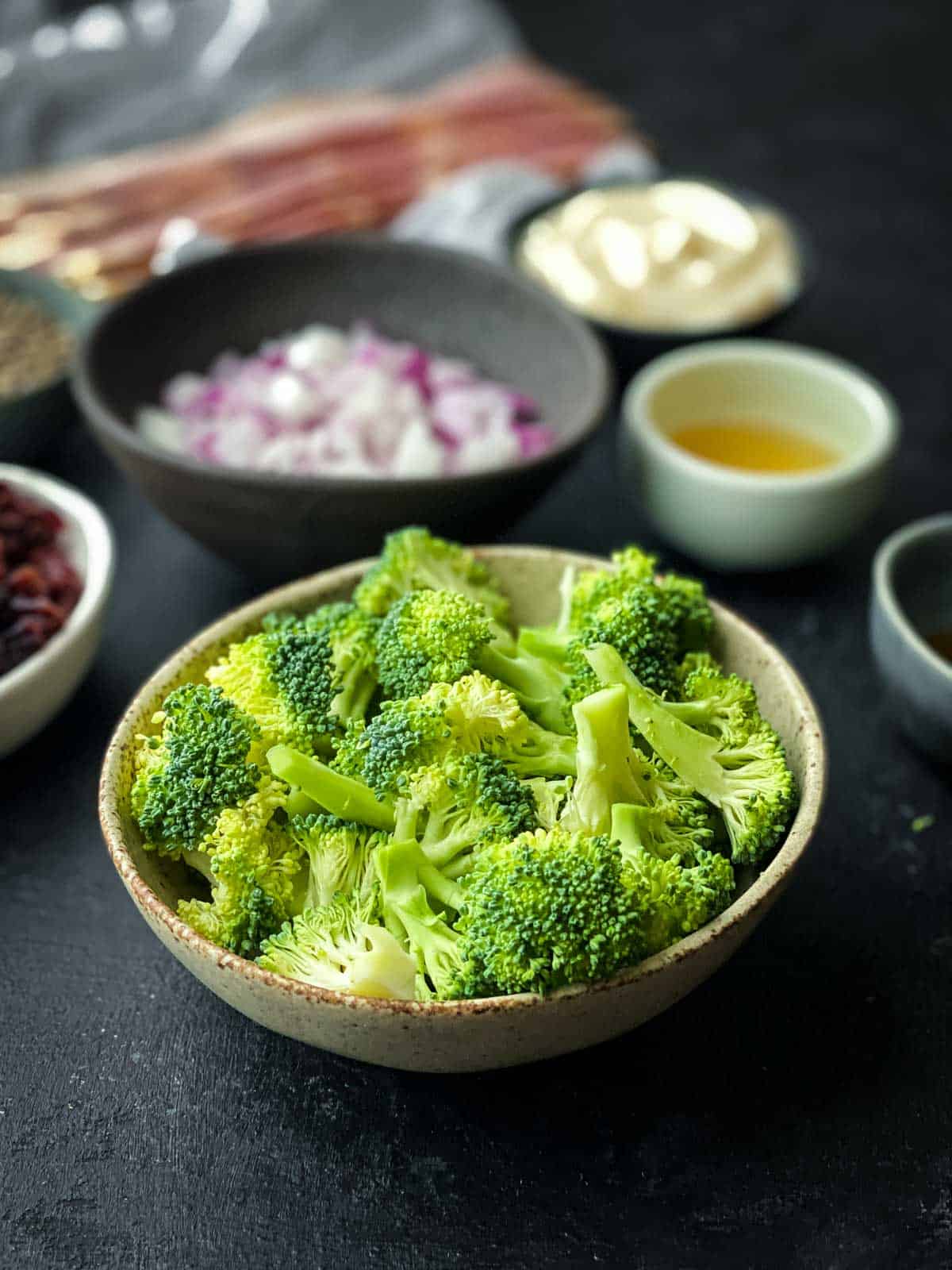 a small bowl of cut broccoli florets in the front with various smaller bowls behind with other broccoli salad ingredients.