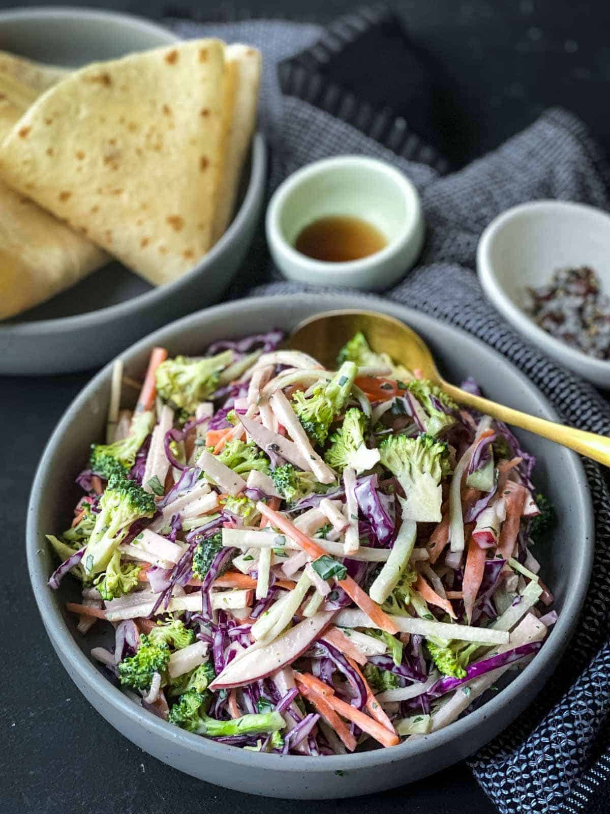 broccoli slaw in a round grey bowl with serving spoon in front with flatbread, syrup and salt and pepper at the back.