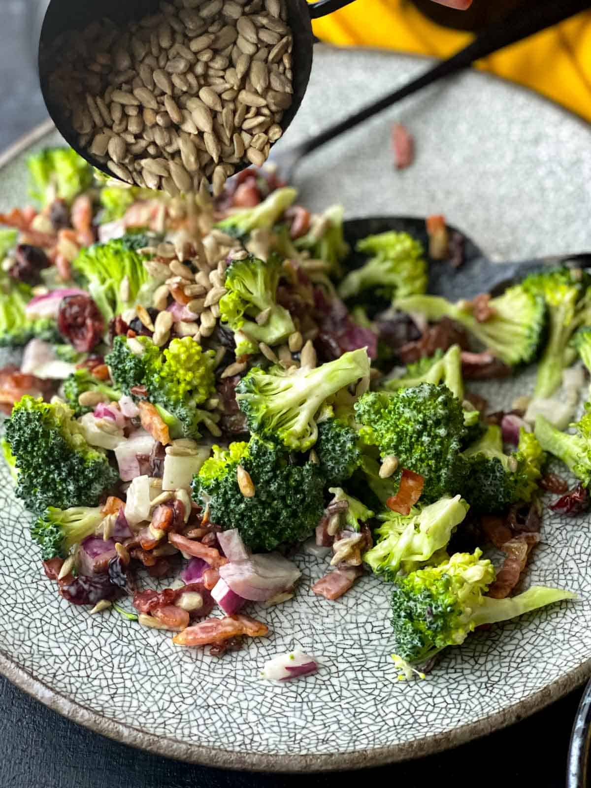 a plate of broccoli salad with sunflower seeds being sprinkled on top