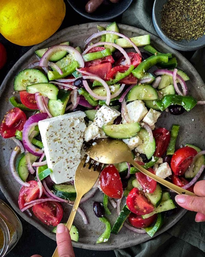 Greek salad with fork and spoon on a plate with small bowls of olives, dried oregano and lemon.