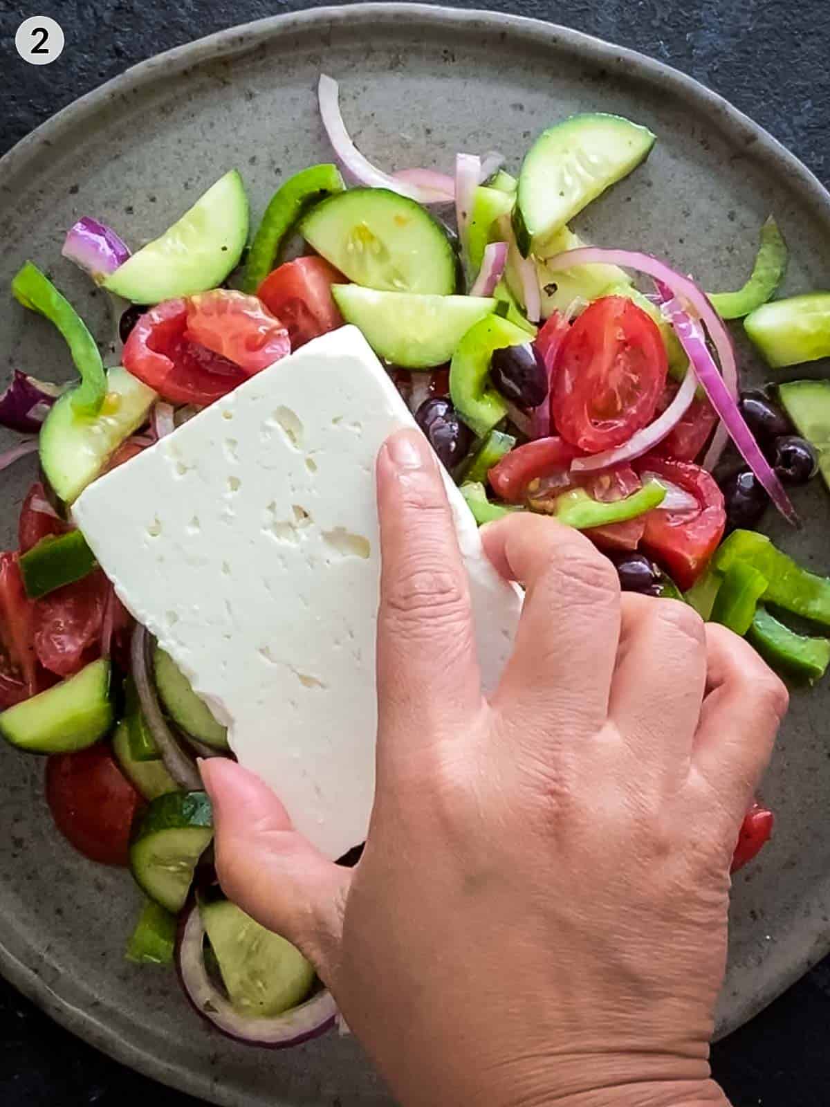A hand placing a block of feta on top of Greek salad on a brown plate