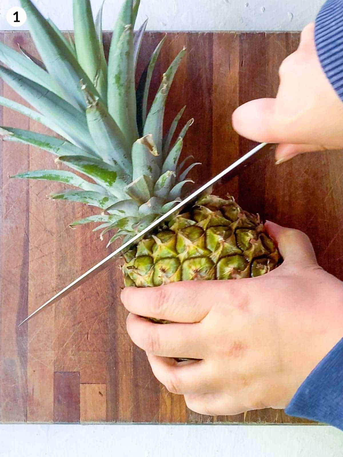 hand holding knife cutting the top of pineapple on a wooden chopping board