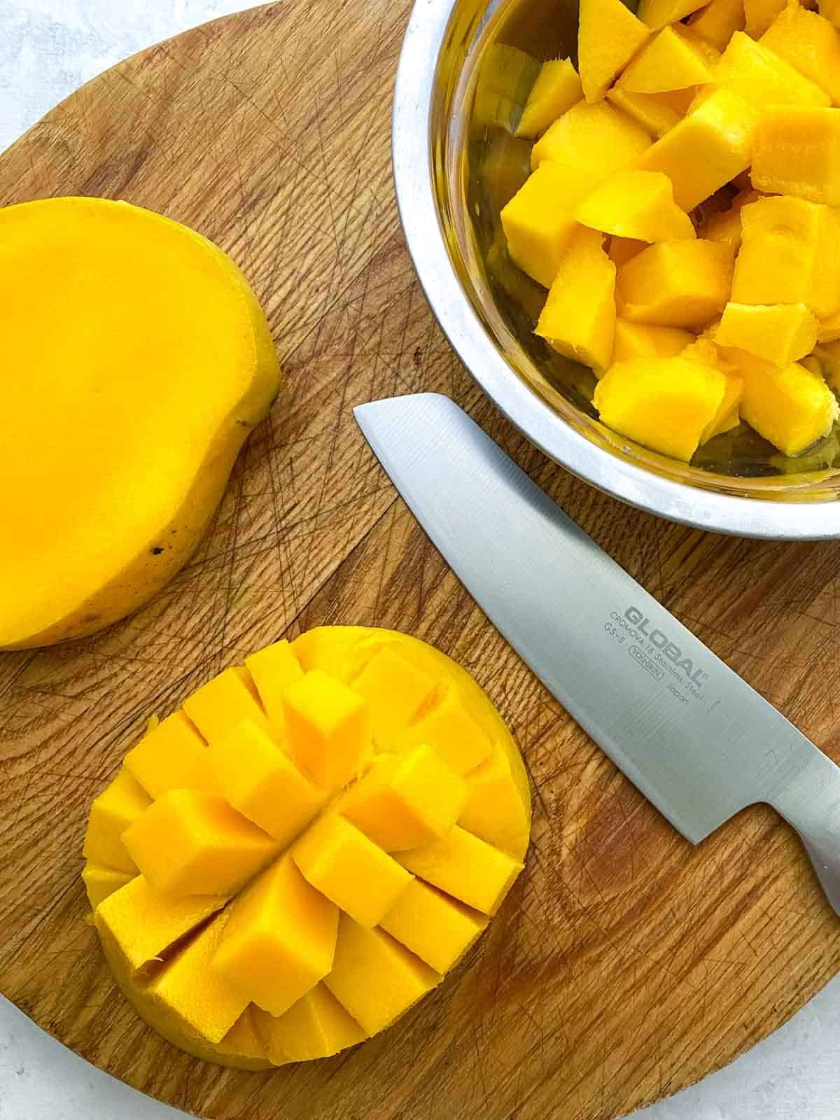 A bowl of cubed mango flesh, half a sliced mango cheek and the mango seed on a wooden board