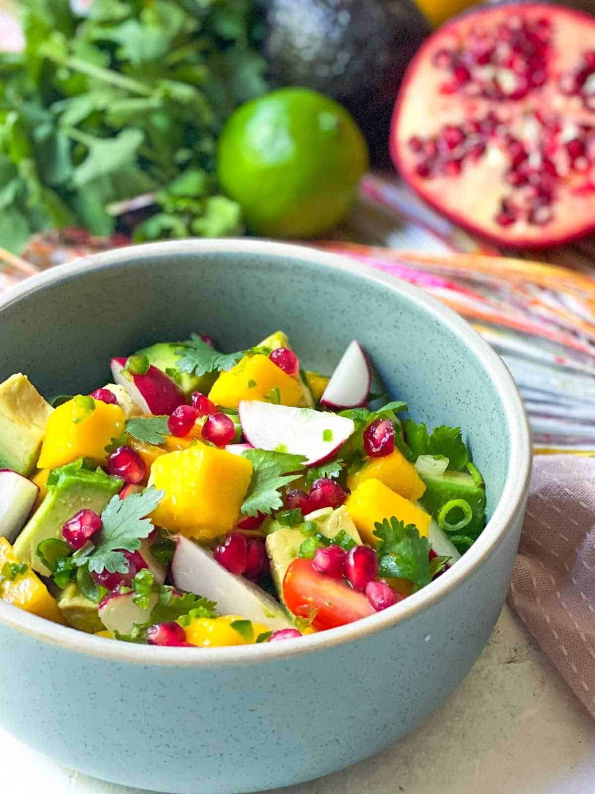 A bowl of Spicy Mango Avocado Salsa with half a pomegranate, whole live, avocado and coriander in the background