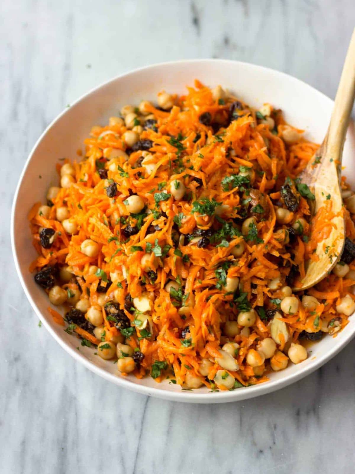 Carrot chickpea salad in a white bowl and a wooden salad spoon on a marble tabletop