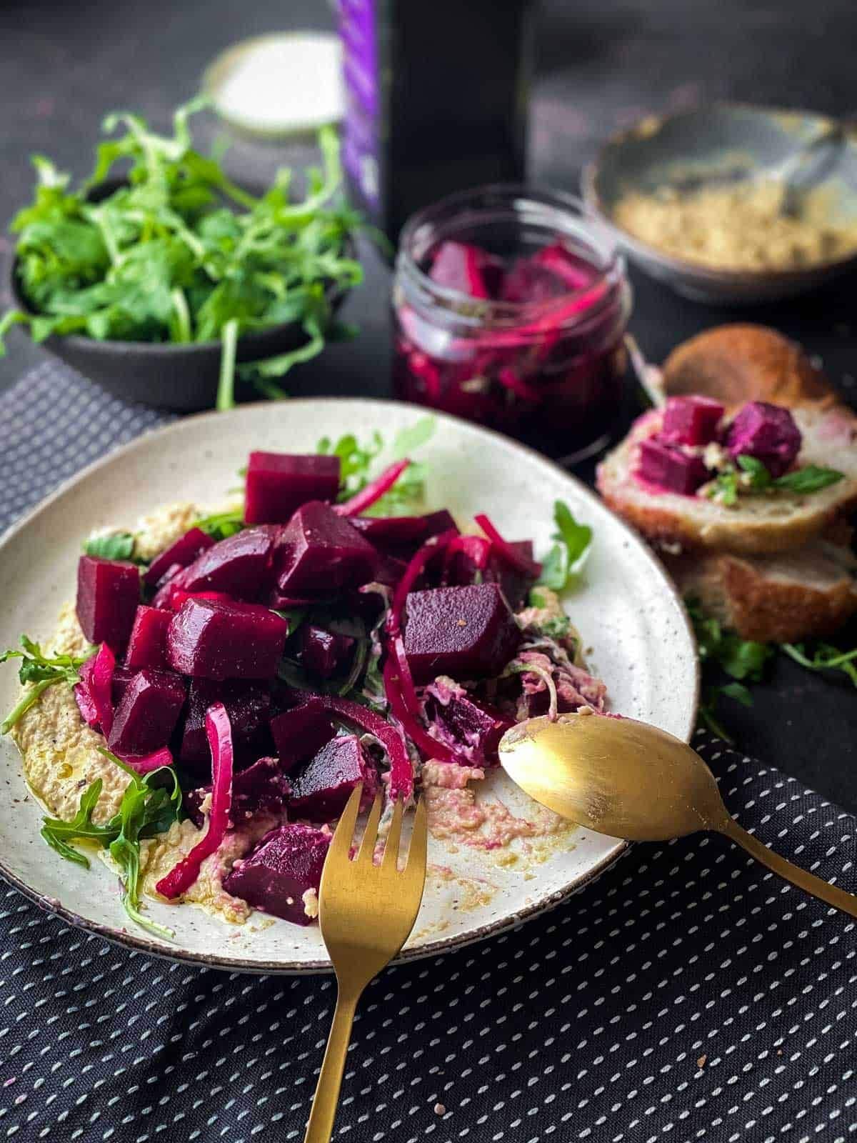 Pickled beetroot salad on chickpea mash served on a white plate and gold cutlery with bread, olive oil, arugula and pickled beets on the side