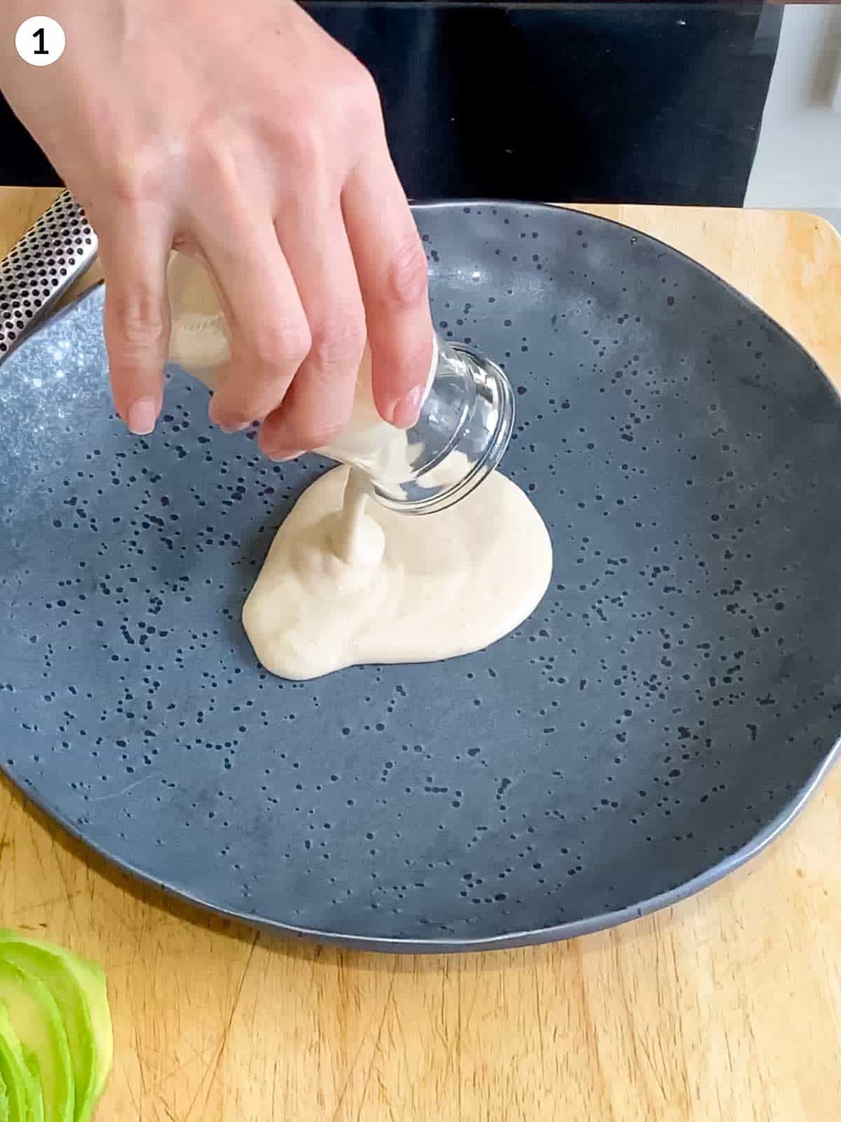 Hand pouring creamy garlic dressing in a glass jar onto a grey plate