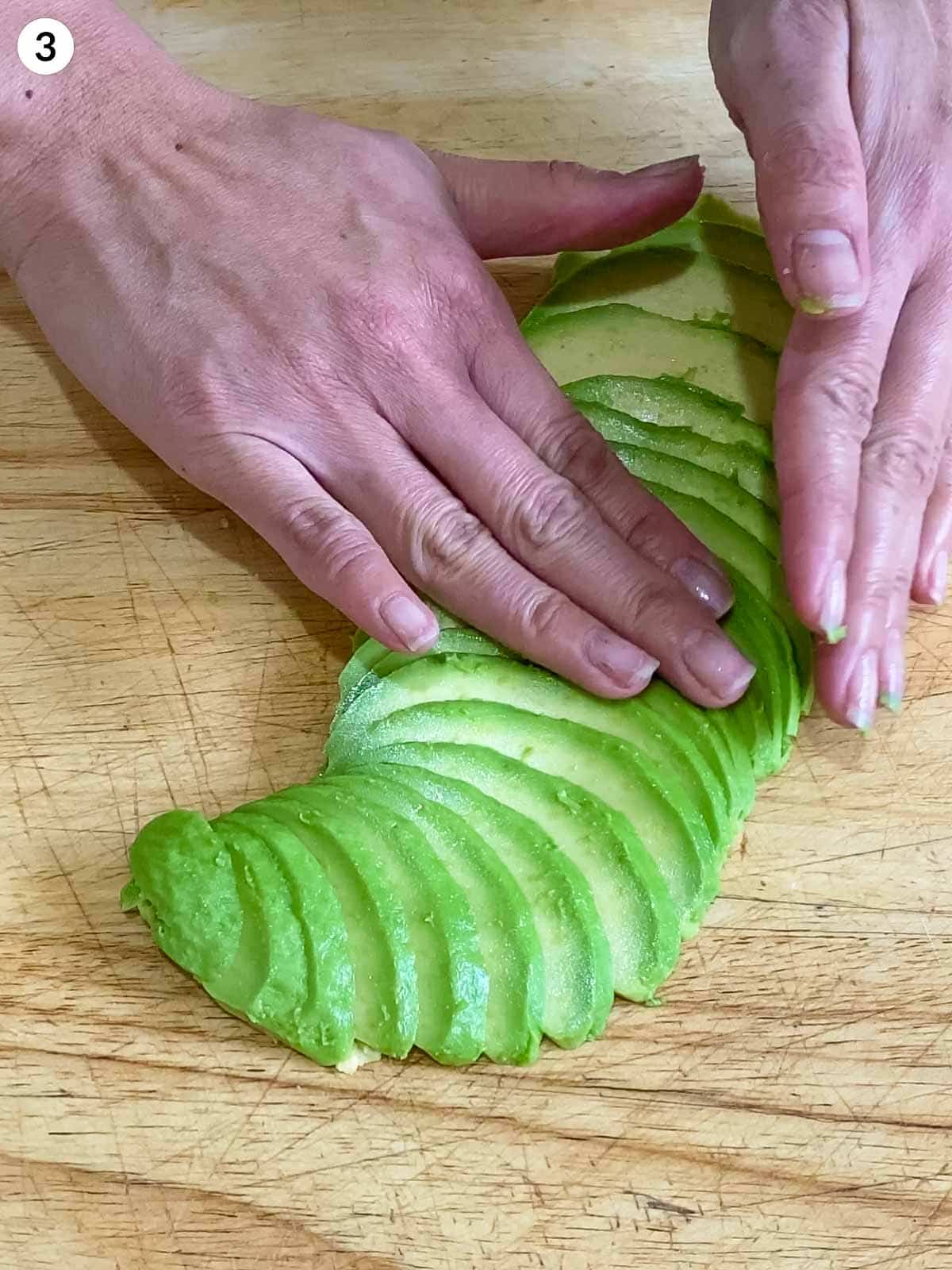 2 hands making avocado fan on a wooden chopping board