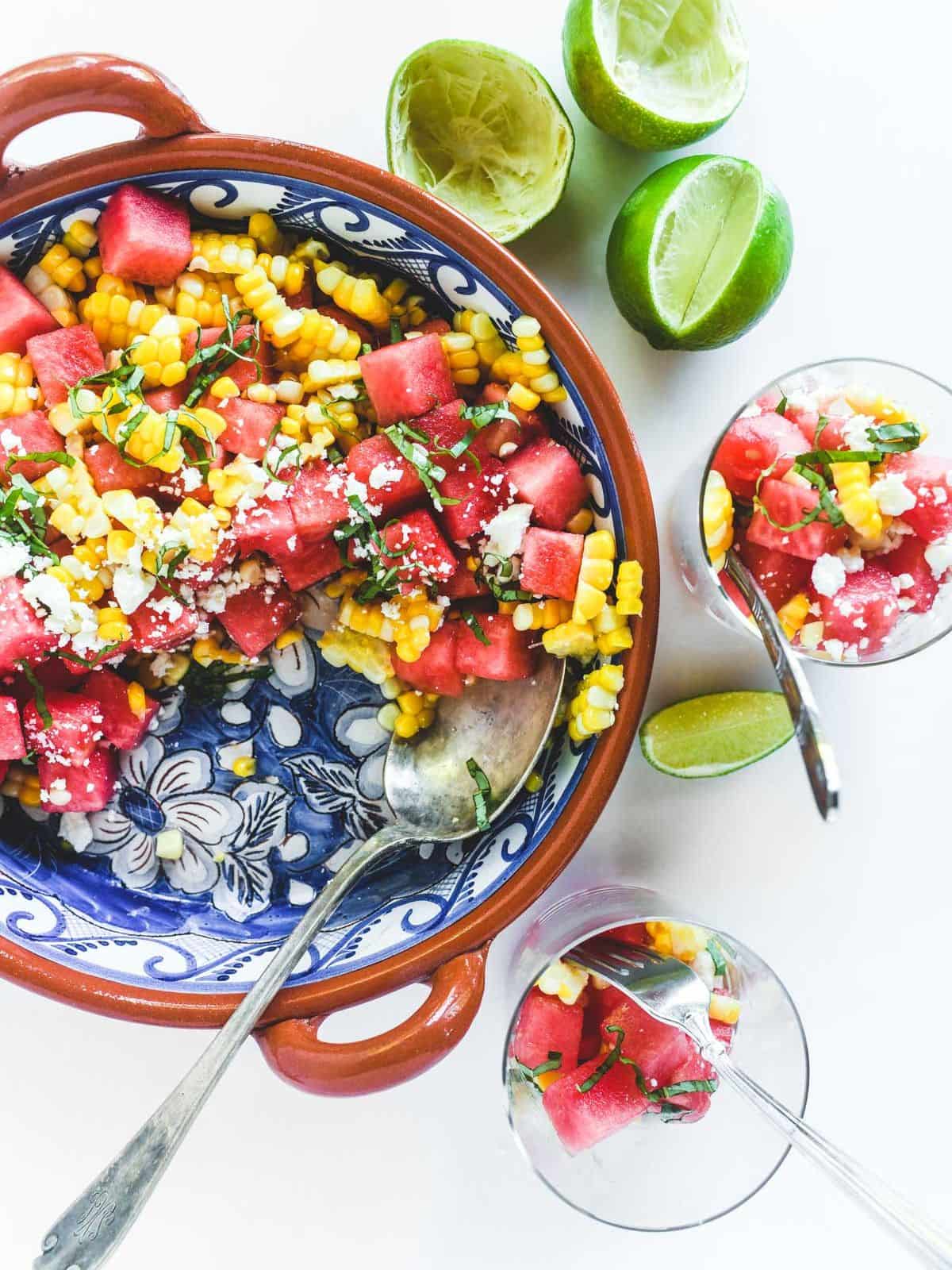 watermelon corn salad in a Spanish bowl with handle, served with half limes on the side a salads in glass cups