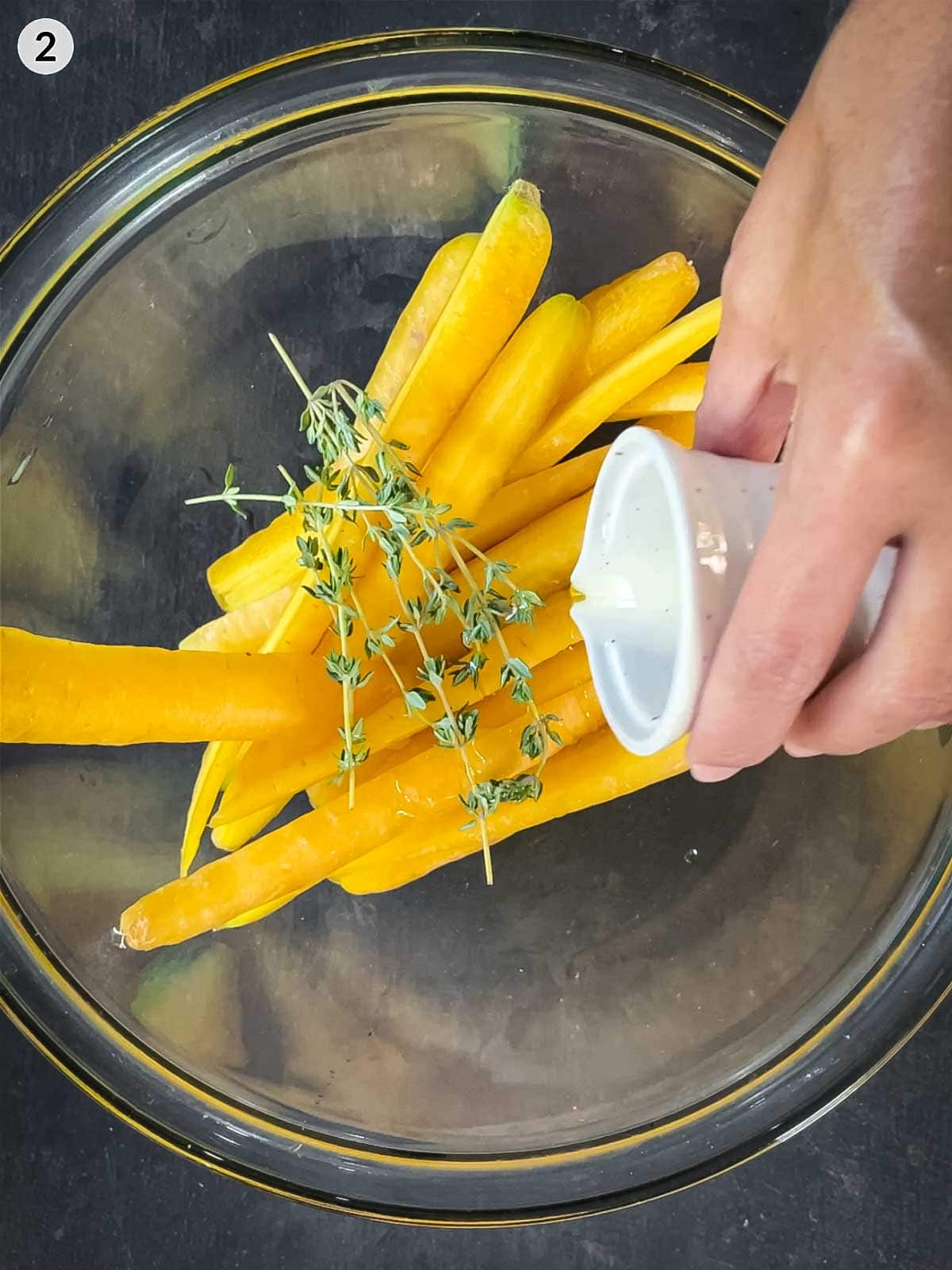Pouring avocado oil form a cermaic jug over yellow carrots and fresh thyme in a glass prep bowl