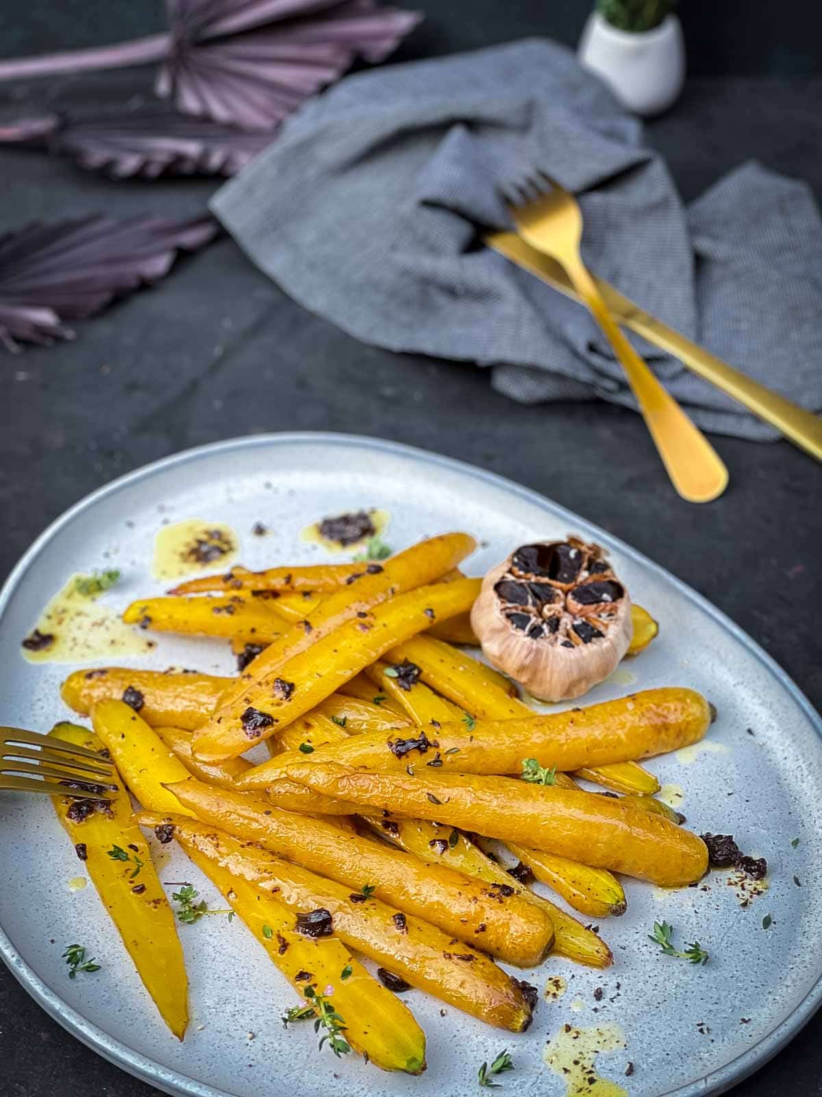 Roasted yellow carrots and black garlic on a blue salad platter with gold cutlery and blue napkin in the background