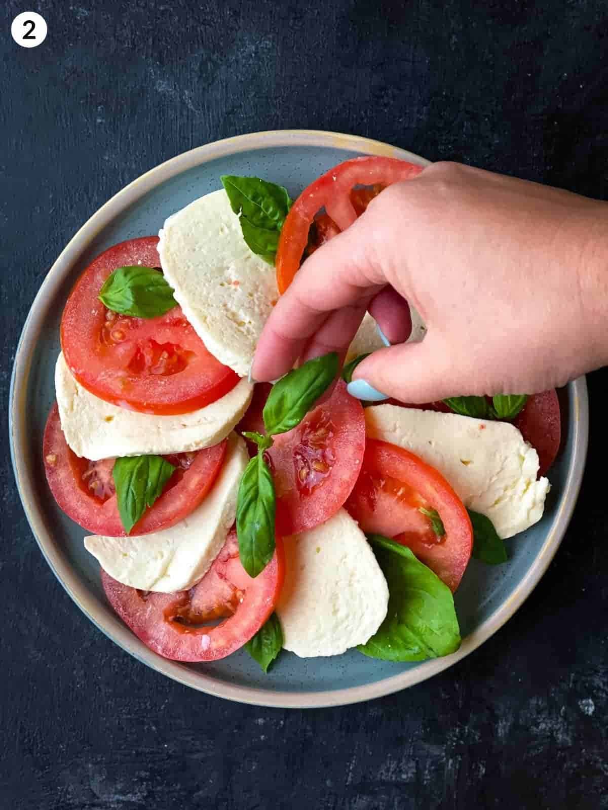 hand placing basil leaf on top of tomato and mozzarella slices on a round plate
