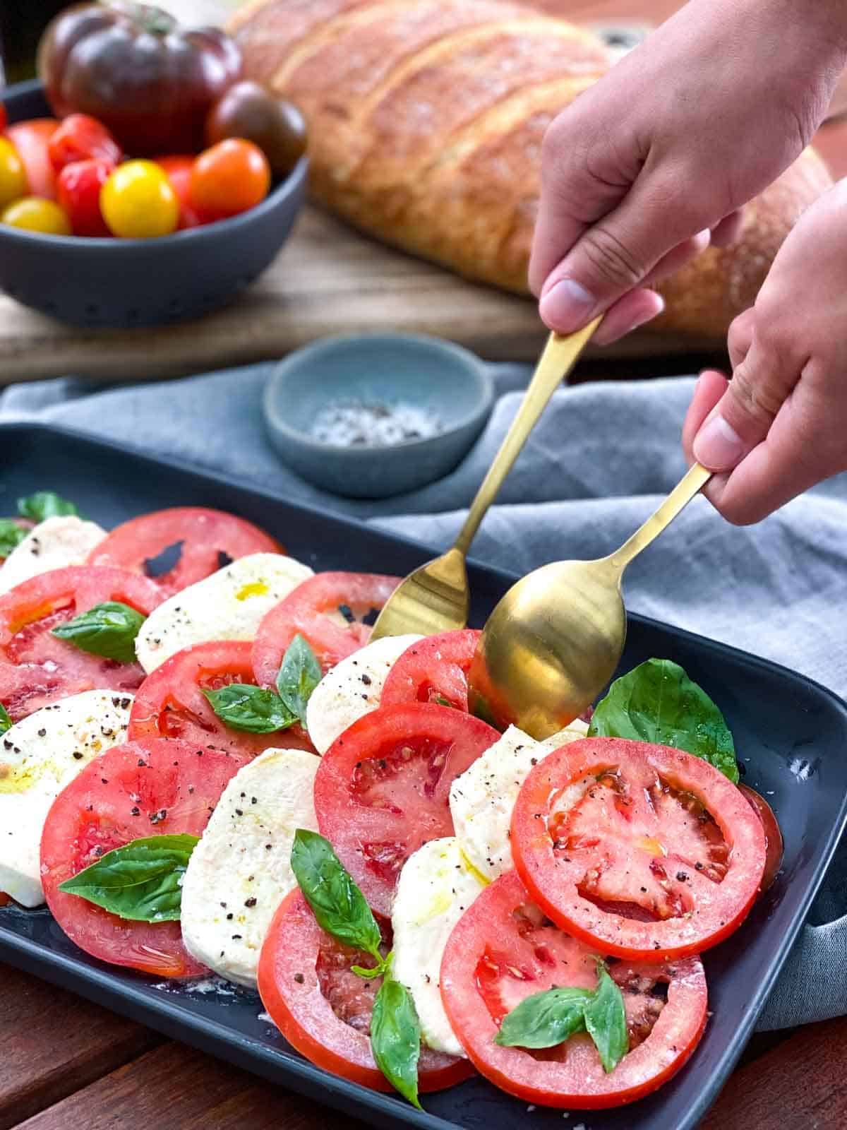 two hands holding fork and spoon to serve caprese salad on a black plate. Bread and bowl of tomatoes in background.