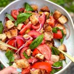 Hand holding Panzanella salad in a white salad bowl with gold cutlery