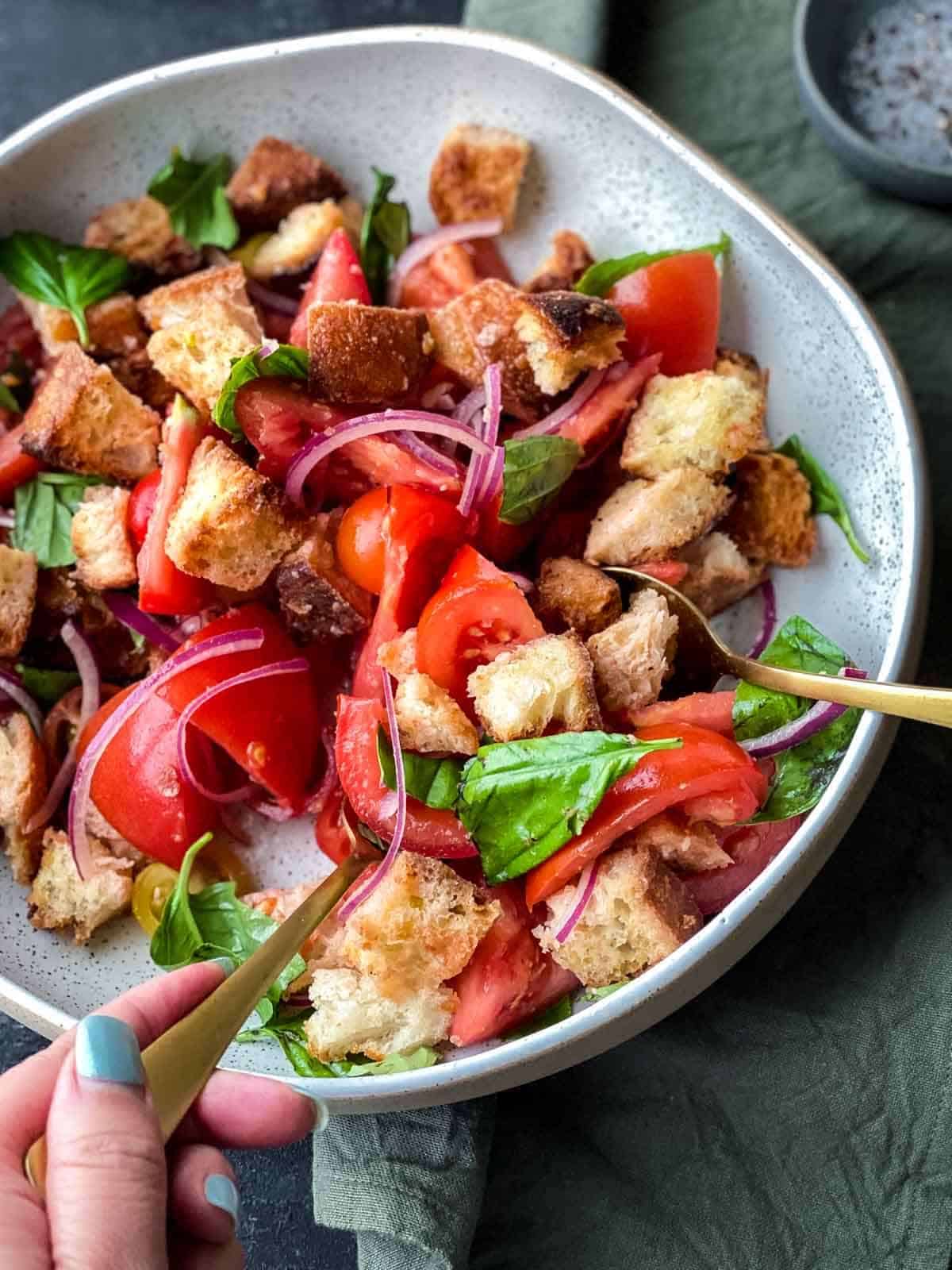 Panzanella salad in a white salad bowl with hand holding gold cutlery