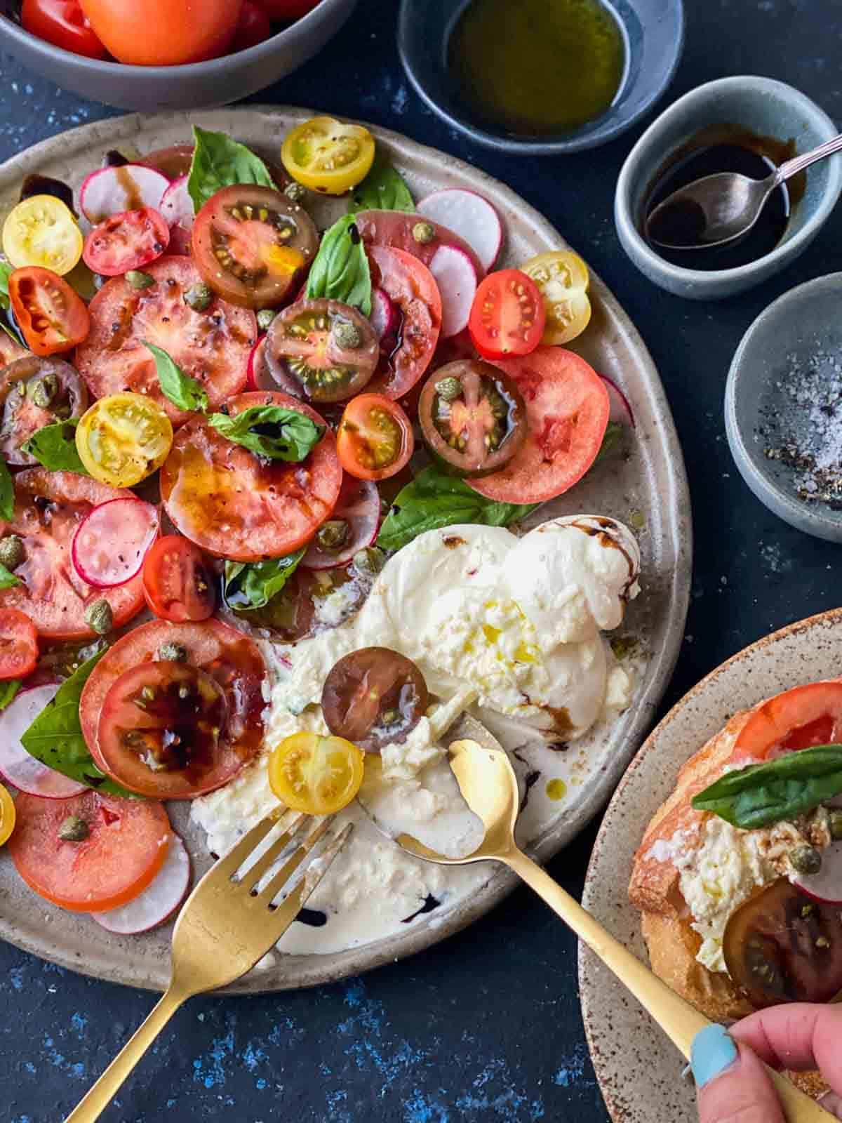 a plate of tomato burrata salad with serving fork and spoon breaking burrata cheese