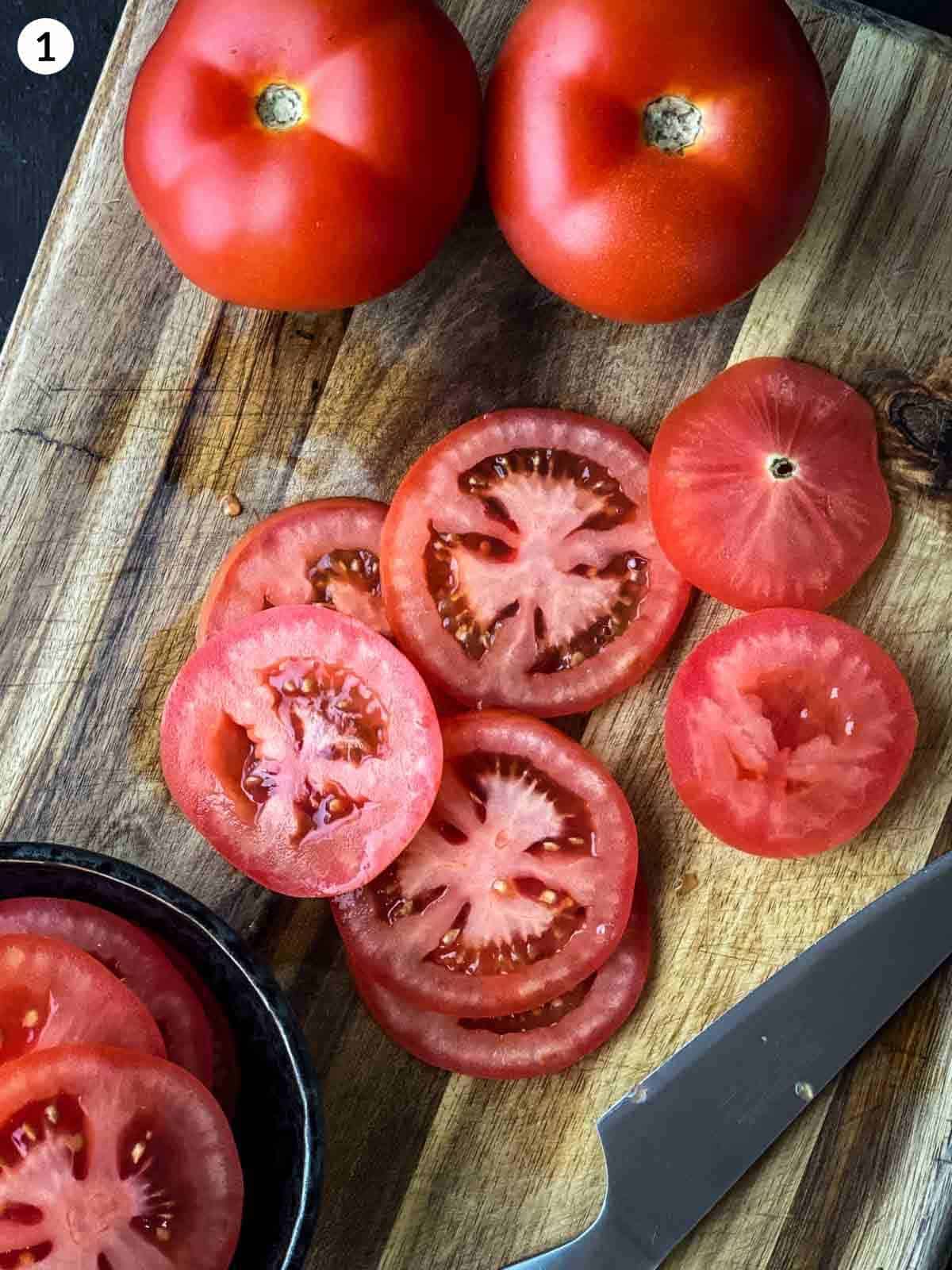 slices of tomato cut on a wooden chopping board with a knife and whole tomatoes next to them.