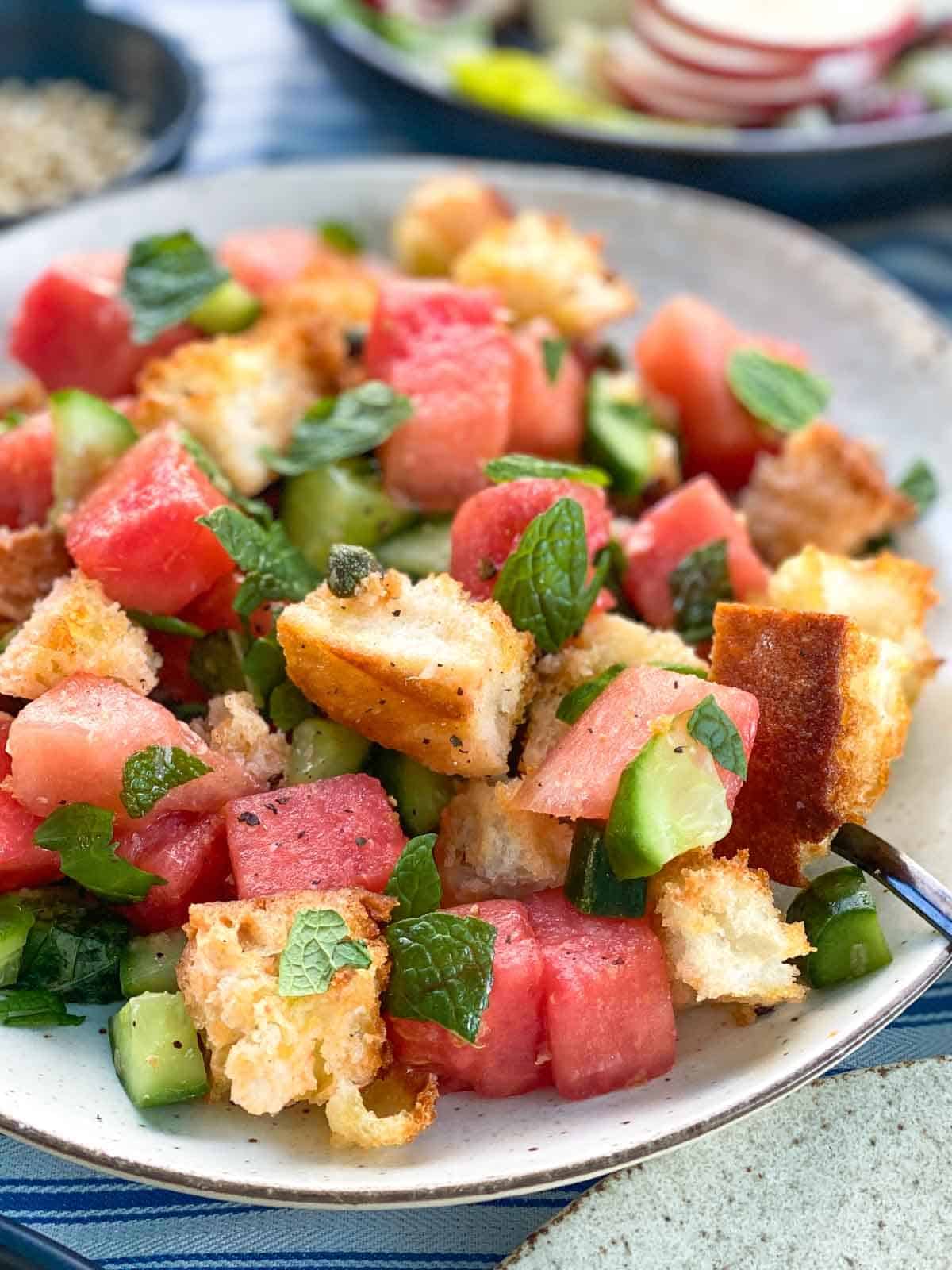 cubes of watermelon tossed with bread, mint, cucumber and capers on a white plate