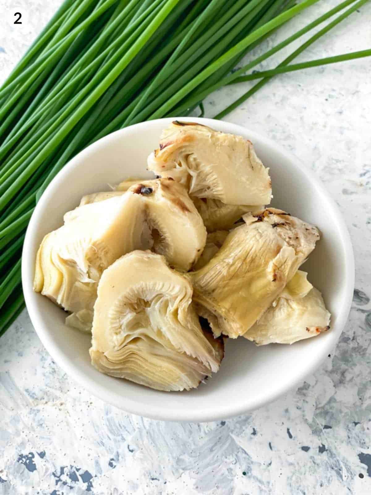 cut marinated artichoke hearts in a small white plate with a bunch of chives placed behind the plate