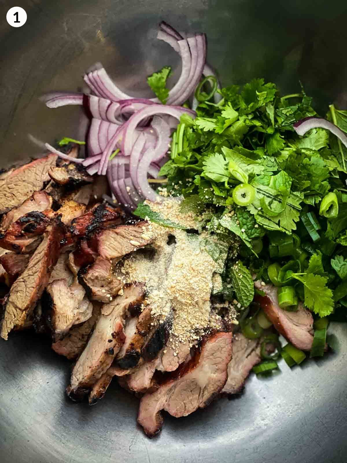 a mixing bowl with sliced grilled pork, mint, cilantro, red onions and toasted rice powder ready to be mixed.