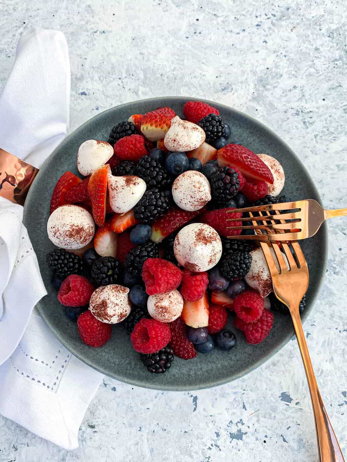 Berry Salad with Sumac Dusted Meringues on a grey plate with 2 gold forks and white napkin on the side
