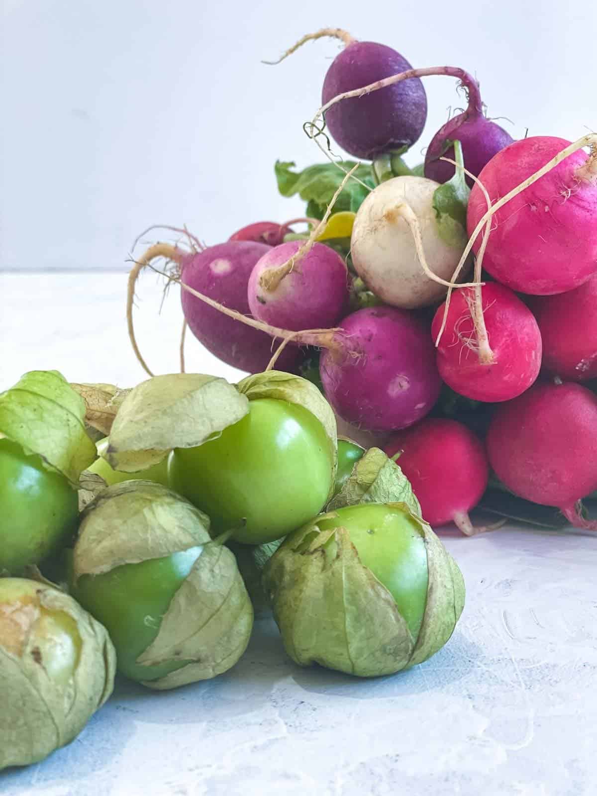 A bunch of rainbow coloured radishes next to whole tomatillos
