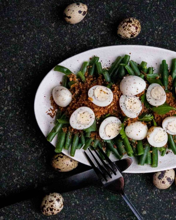Green Bean, Truffle Butter Breadcrumbs and Quail Egg Salad on a white plate with black cutlery and whole quail eggs on the side