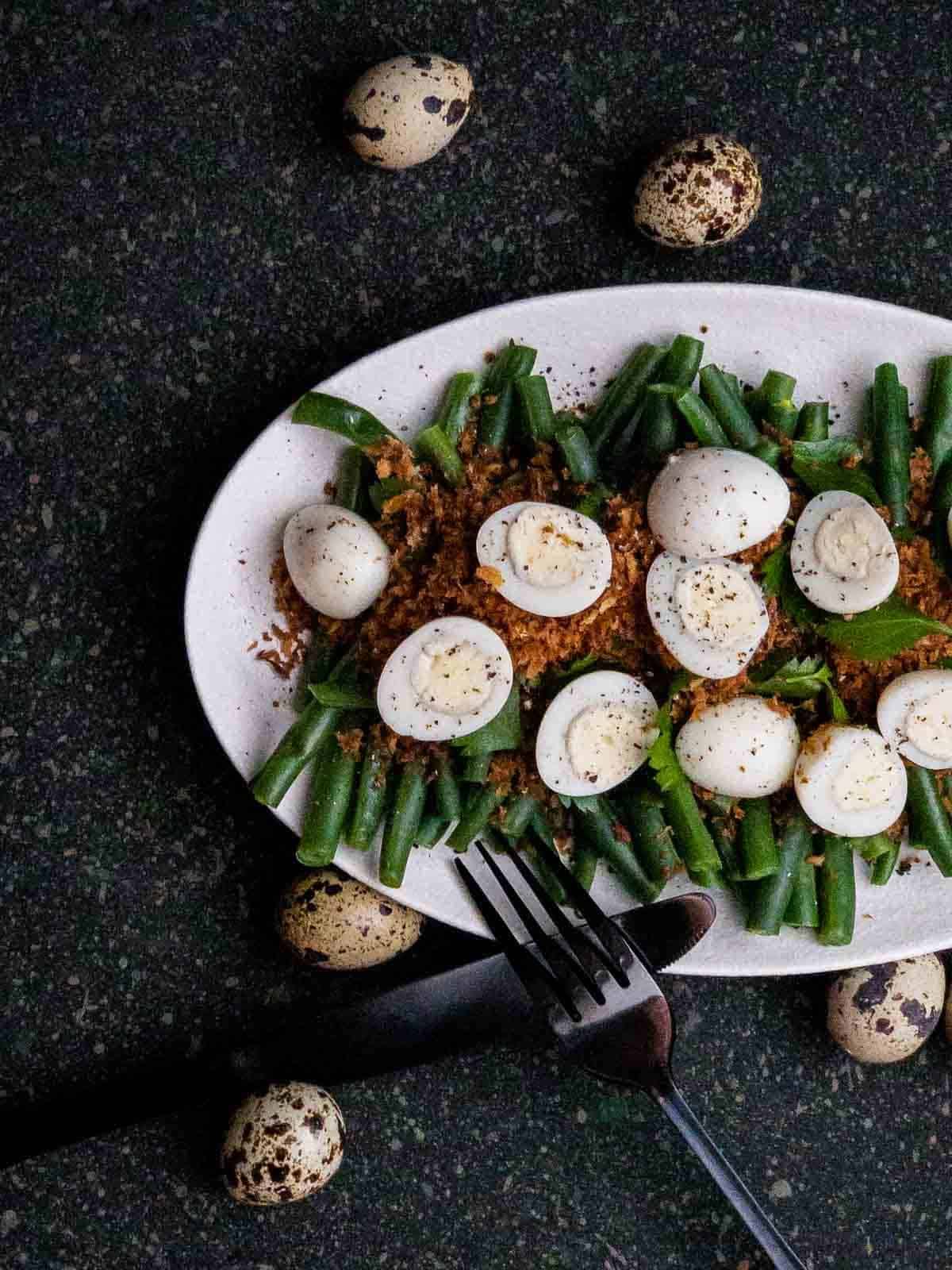 Green Bean, Truffle Butter Breadcrumbs and Quail Egg Salad on a white plate with black cutlery and whole quail eggs on the side