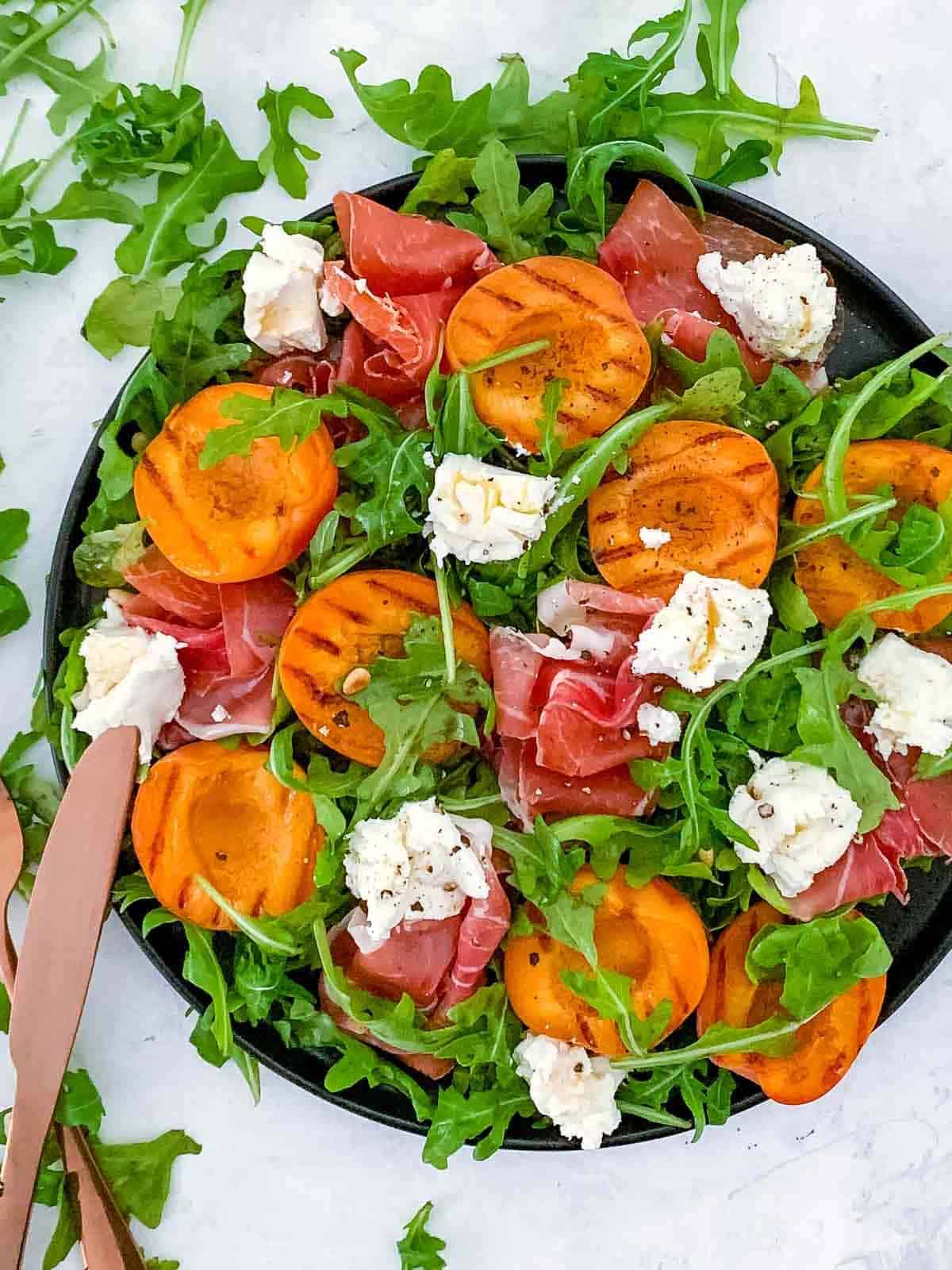 Grilled Apricot Salad with Prosciutto on a black plate with rose gold cutlery