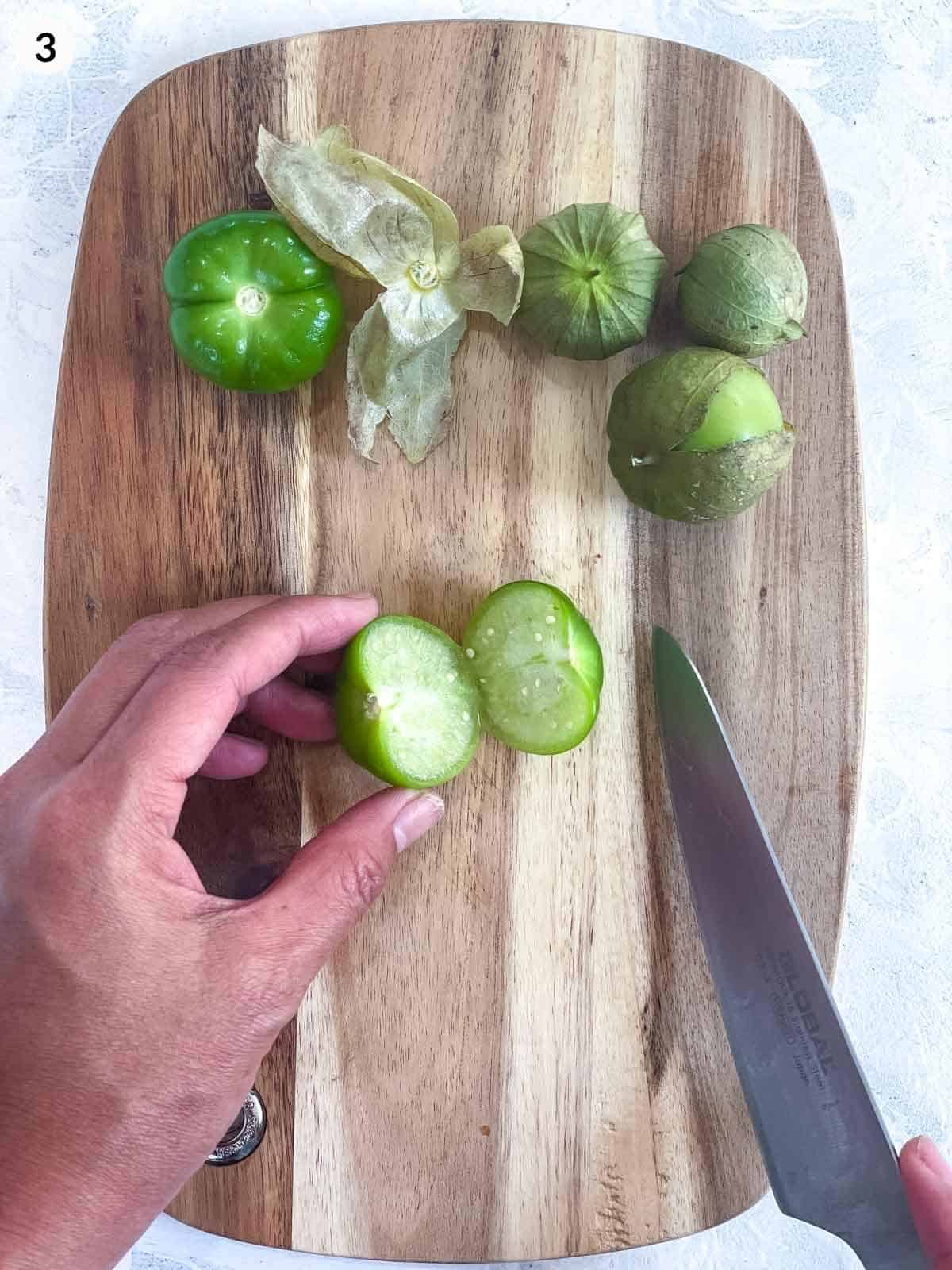 cutting a tomatillo in half with a knife on a wooden board