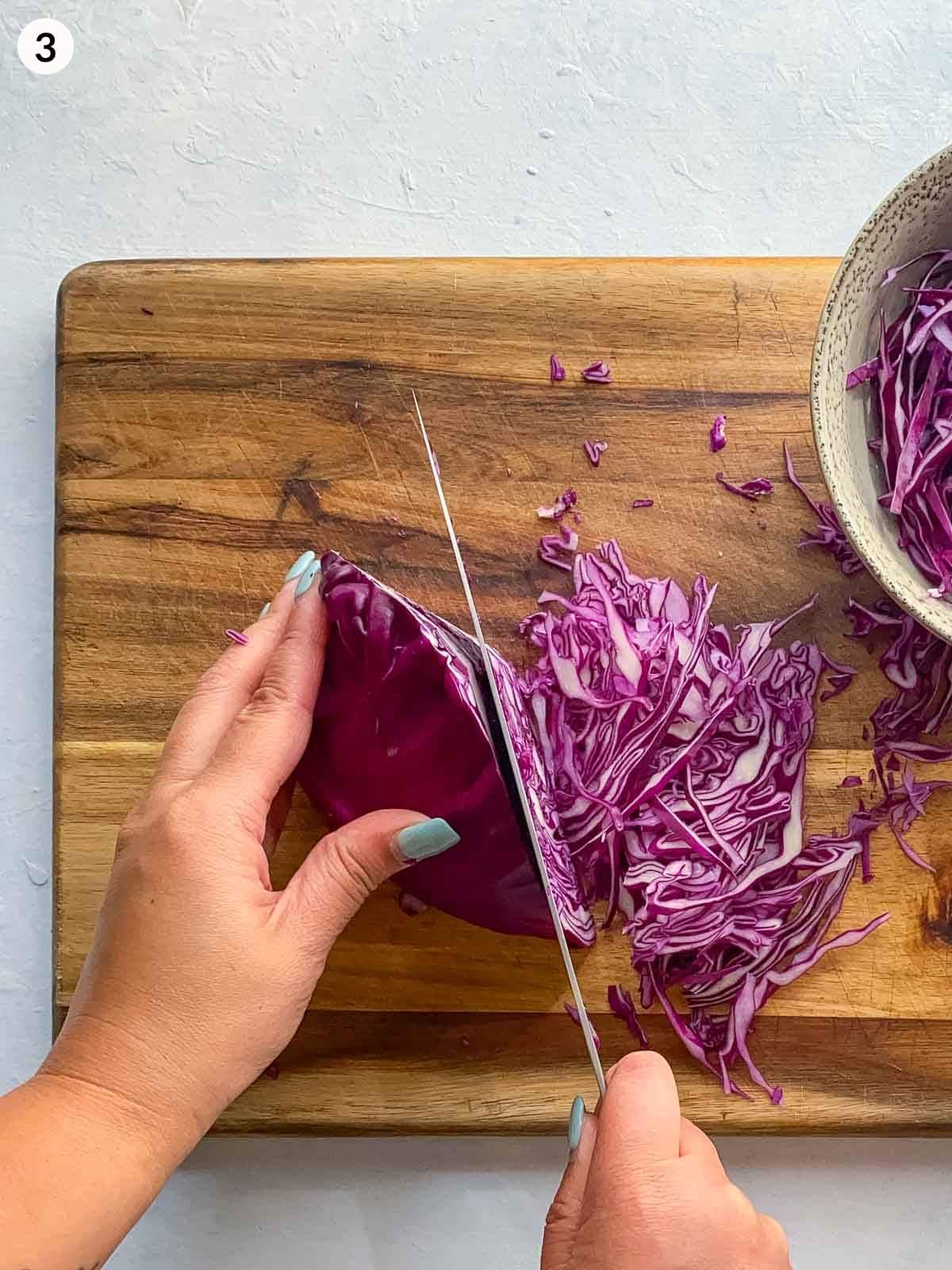 Slicing red cabbage with a knife on a wooden chopping board with a bowl of cabbage on the side