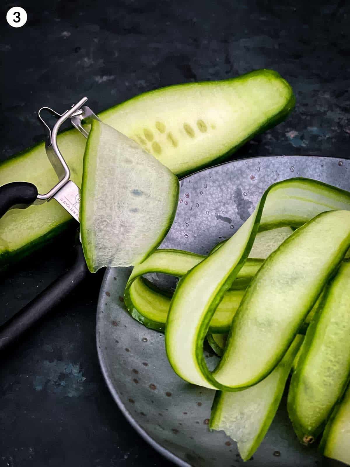 a peeler, peeling side ribbons of cucumber on a grey plate