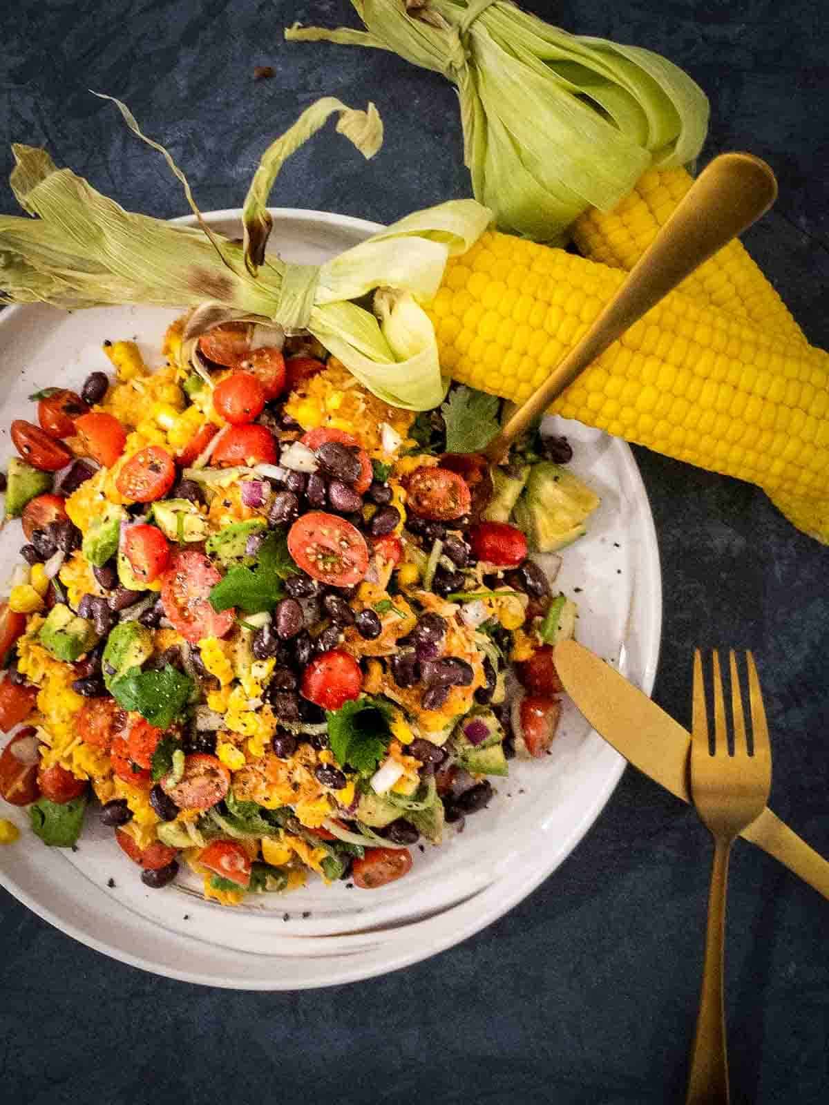 Parmesan Chipotle Corn Salad with Black Beans on a white plate with gold cutlery and 2 corn on the cobs
