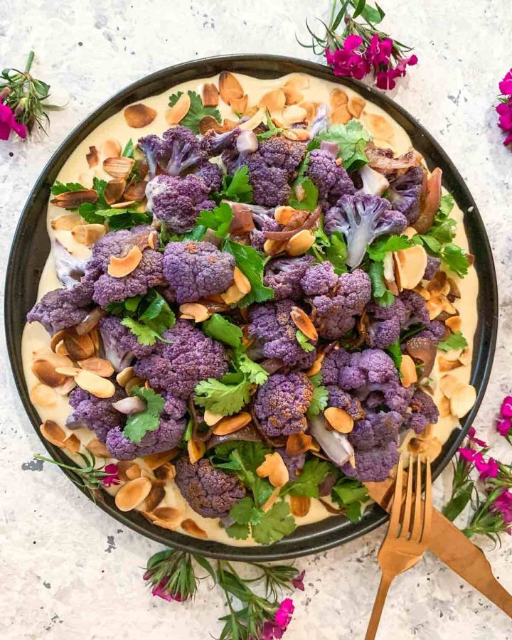 Purple Cauliflower Salad with Lemon Mustard Tahini on a black plate with gold cutlery and pink flowers scattered around the plate