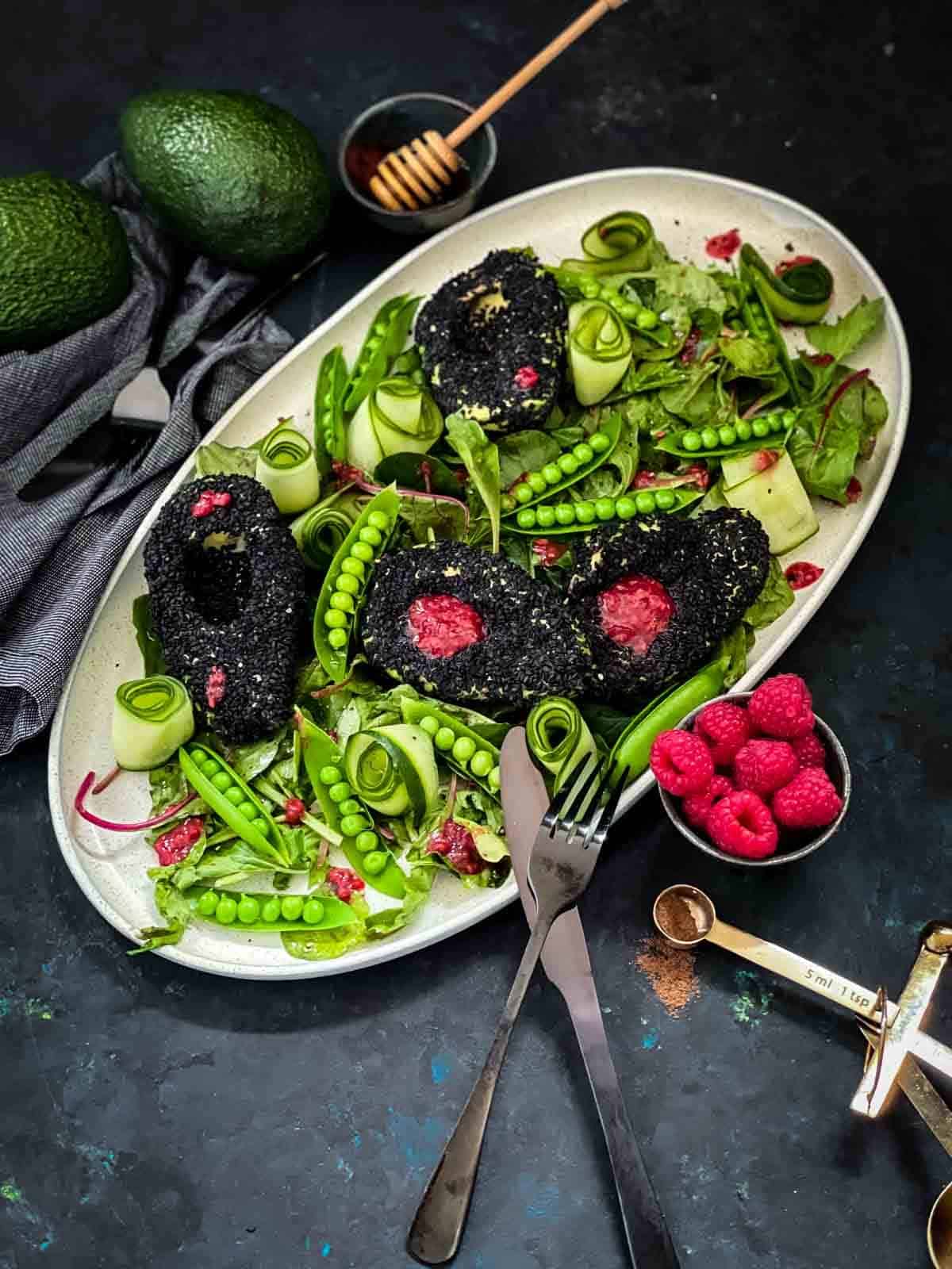 Sesame Avocado Cucumber Salad in a oval salad platter with black cutlery, bowl of fresh strawberries and 2 whole avocados on the side