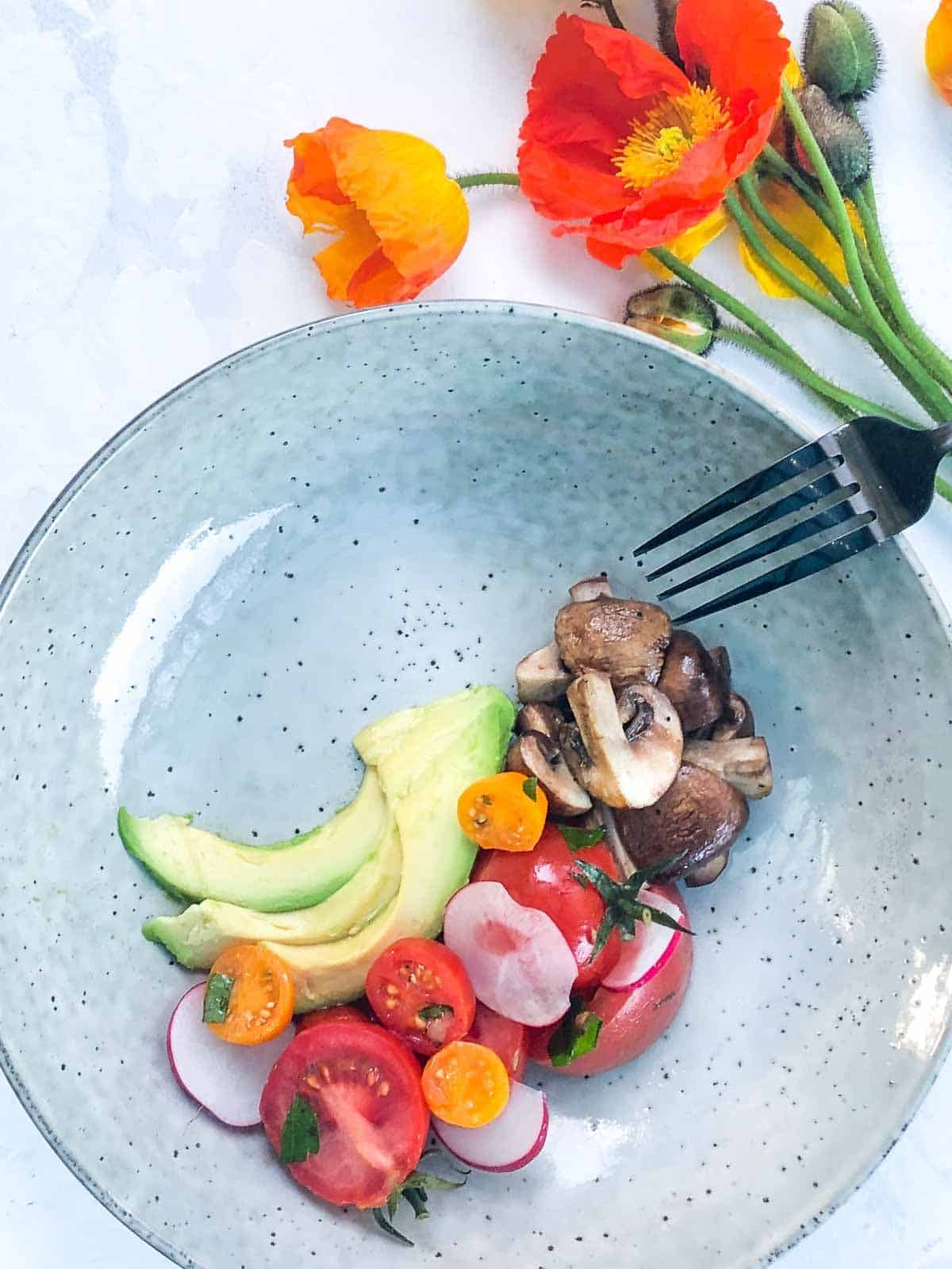 Breakfast Salad with Mushrooms, Avocado and Tomatoes on a blue plate with a black fork and flowers on the side