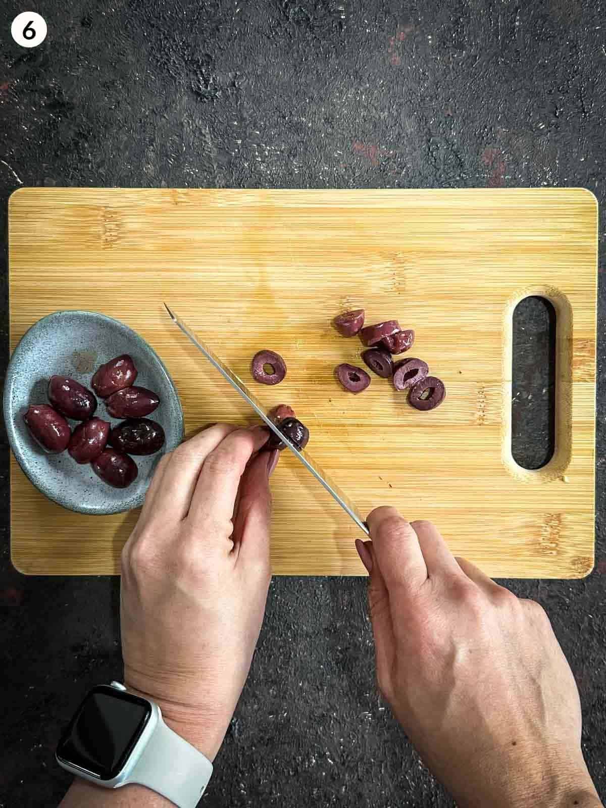 Cutting black olives with a knife on a wooden chopping board with a board of whole olives on the side