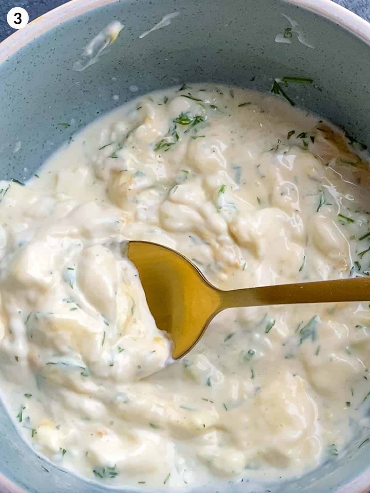 a gold spoon scooping out some creamy salad dressing with dill from a blue bowl