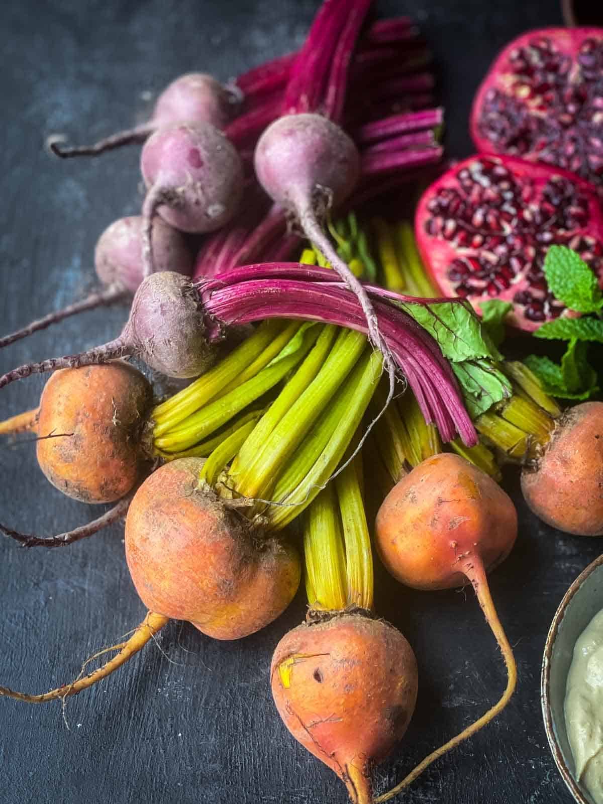 2 bunches of baby beets laid on a black background next to cut pomegranate