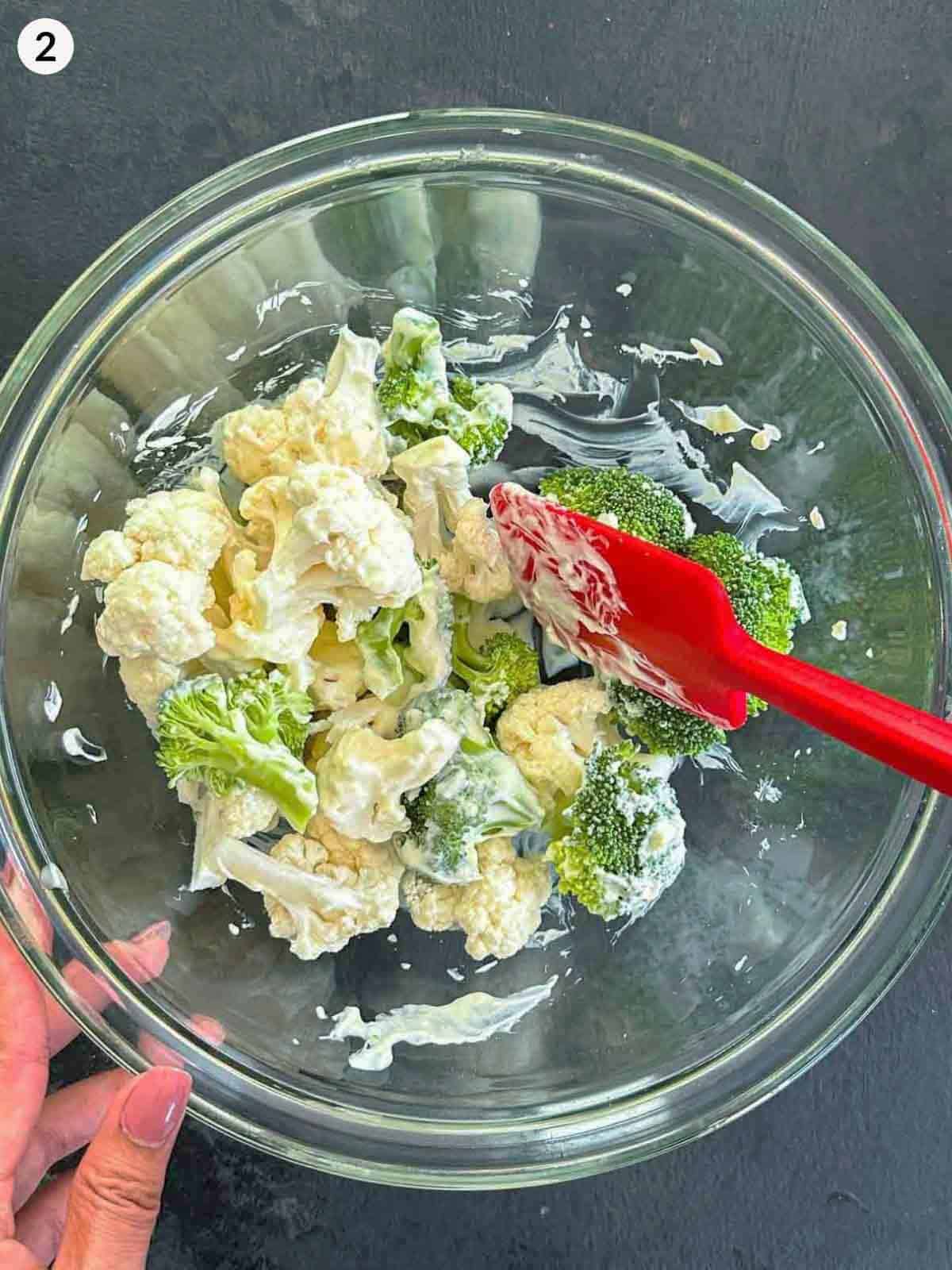 Mixing broccoli and cauliflower florets with mayonnaise in a glass bowl with a red spatula