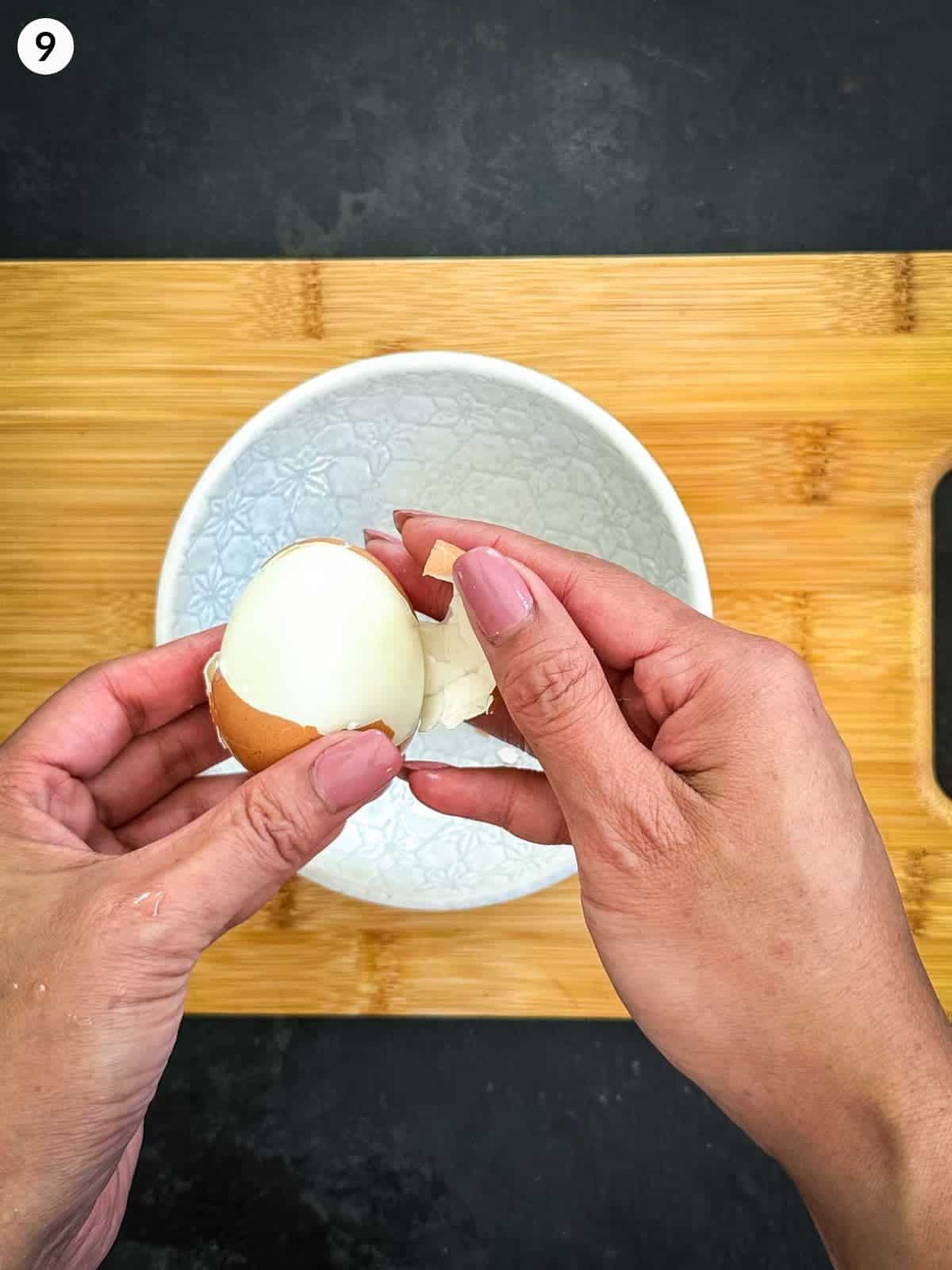 Peeling hard boil egg into a white bowl on a wooden chopping board