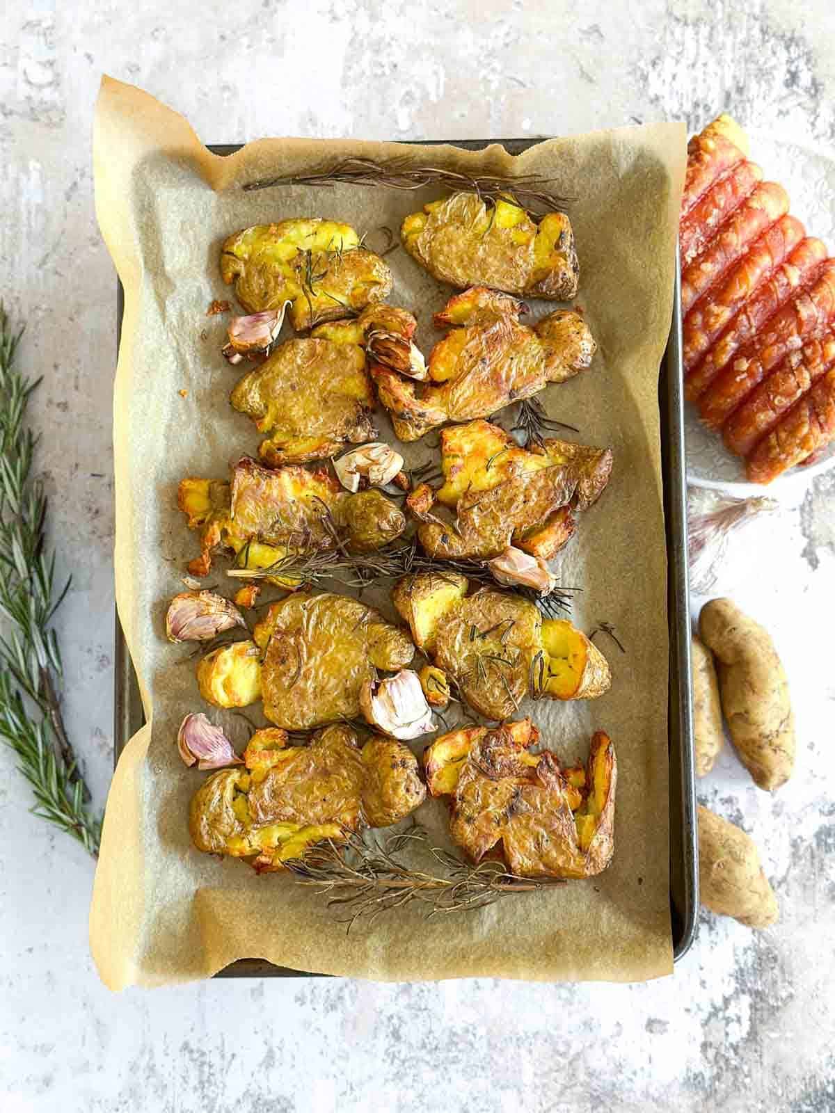 Sheet pan of smashed fingerling potatoes on parchment paper with roast pork, garlic, fresh potatoes and rosemary in the background.