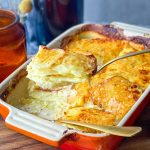 Lifting a serve of Creamy Potatoes au Gratin from a ceramic baking dish with carrots and roast chicken on the side