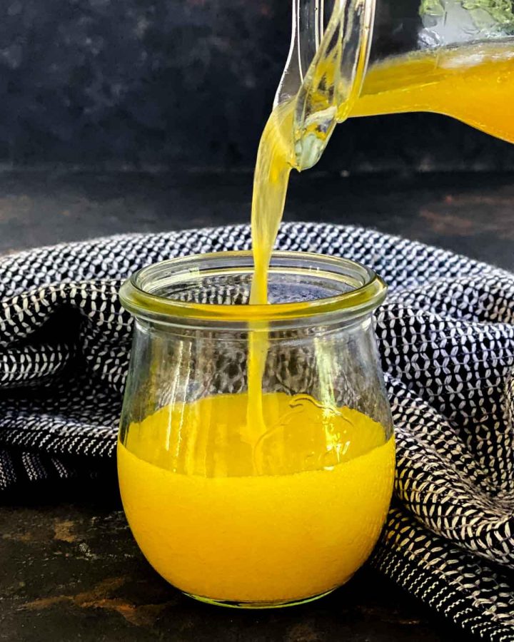 Pouring Apple Cider Vinaigrette with Orange Juice from a jug into a small jar with blue linen in the background